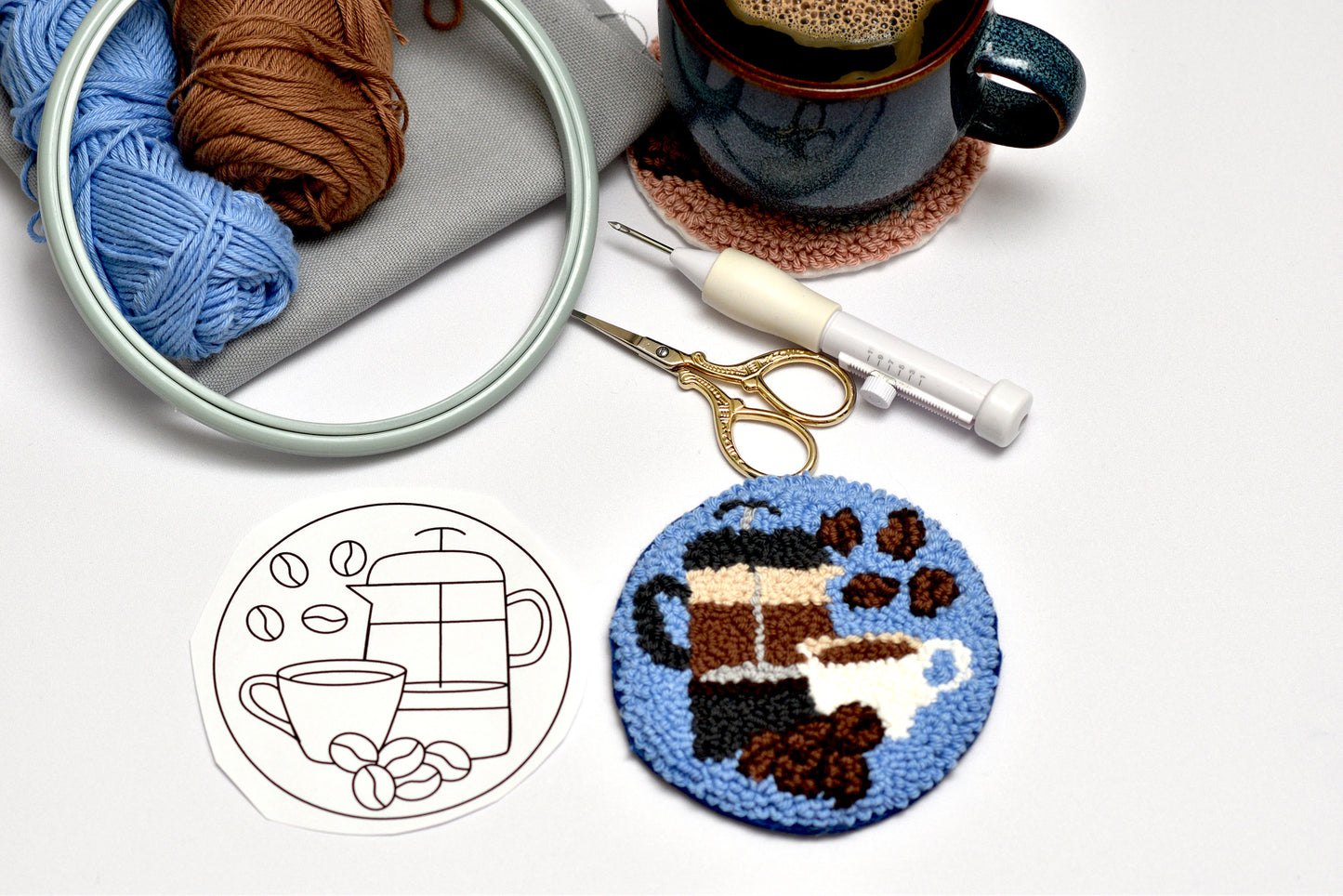 Embroidery hoop with yarn, a cup of coffee, and an embroidered coaster on a white background.