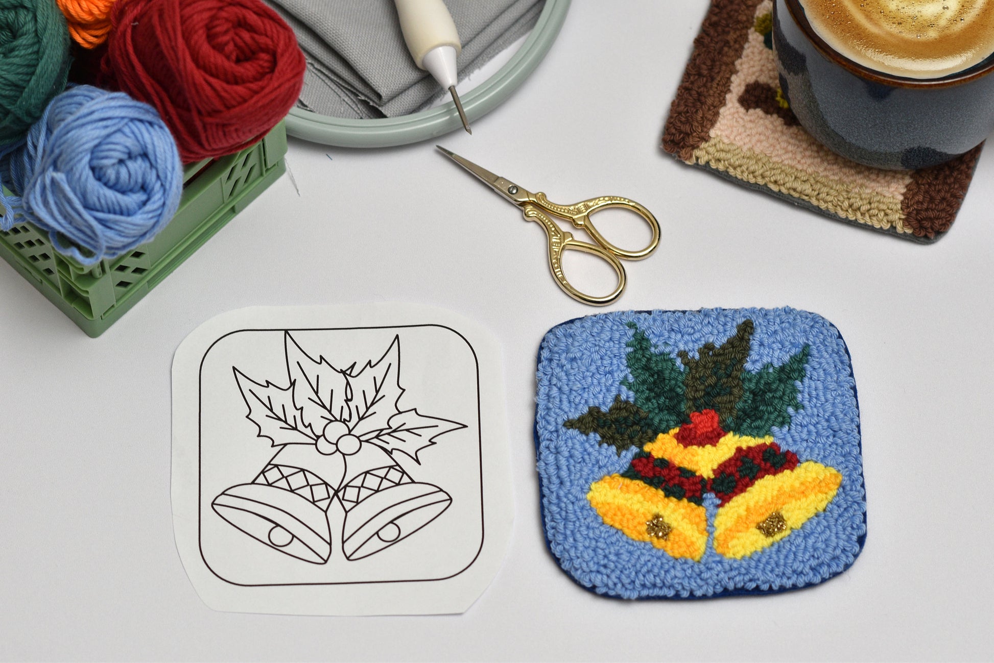 Punch needle coaster with holly design next to a black and white outline, scissors, and yarn on a table.
