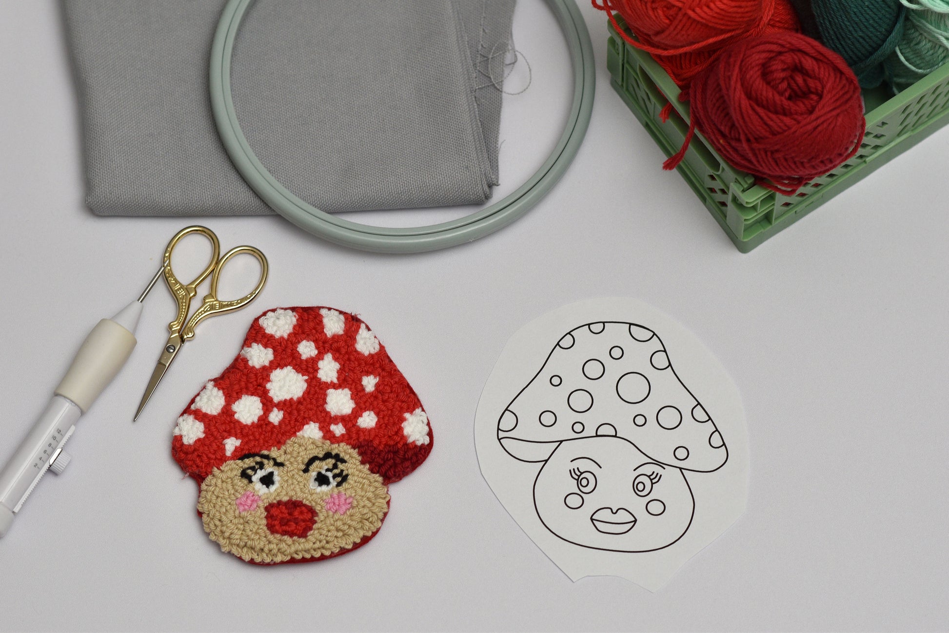 Embroidery hoop, scissors, and red mushroom-shaped embroidered coaster and its design on a white surface.