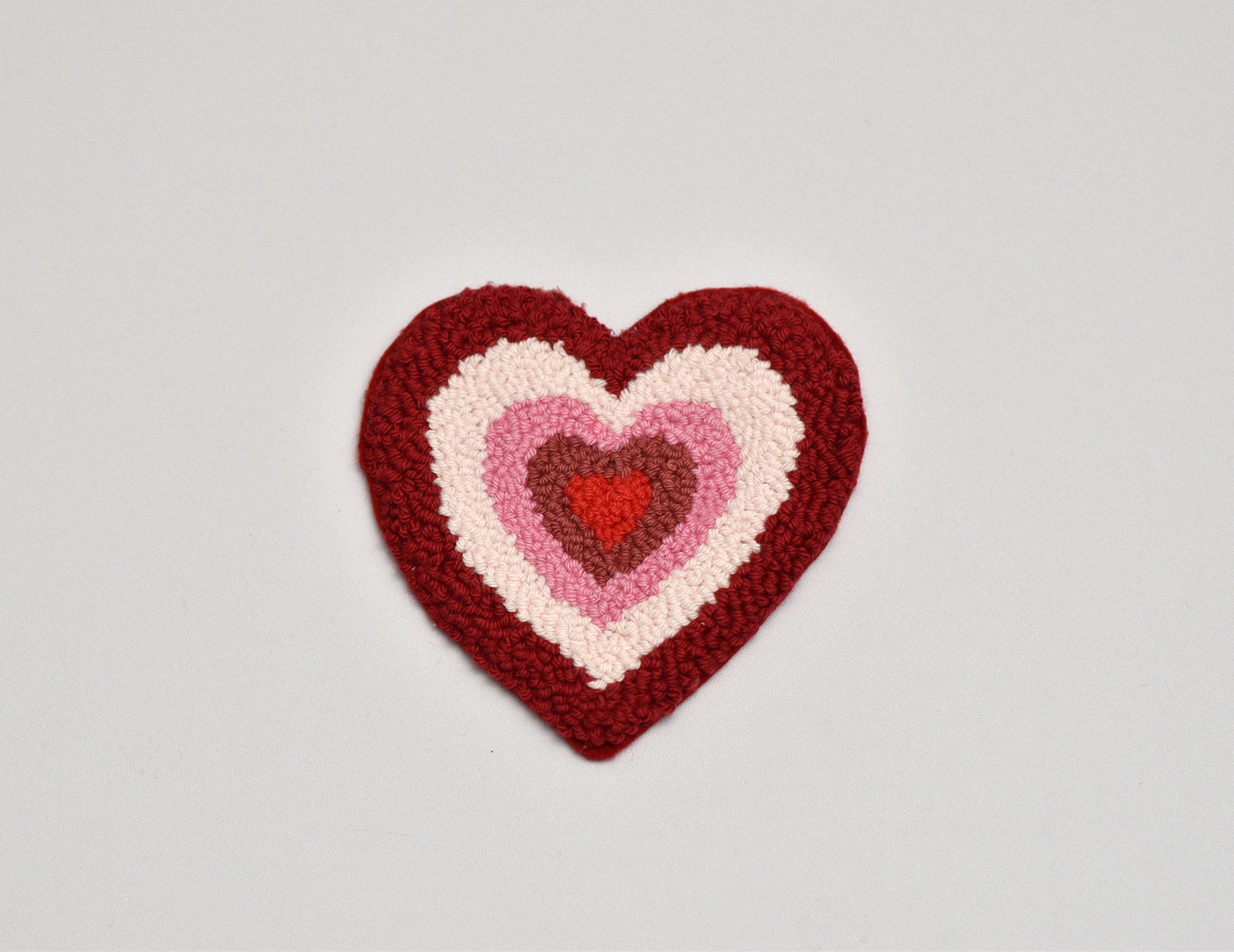 Heart-shaped knitted patch with three concentric hearts in red, pink, and white on a light gray background.
