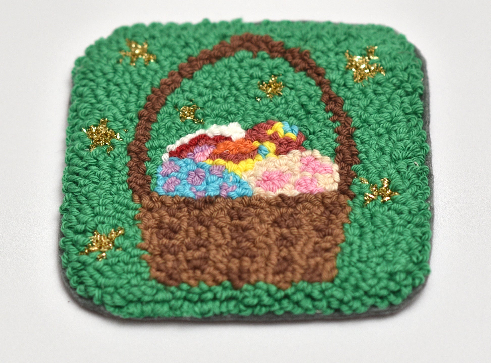 Embroidered coaster of a brown basket full of Easter Eggs with colorful flowers on a green background