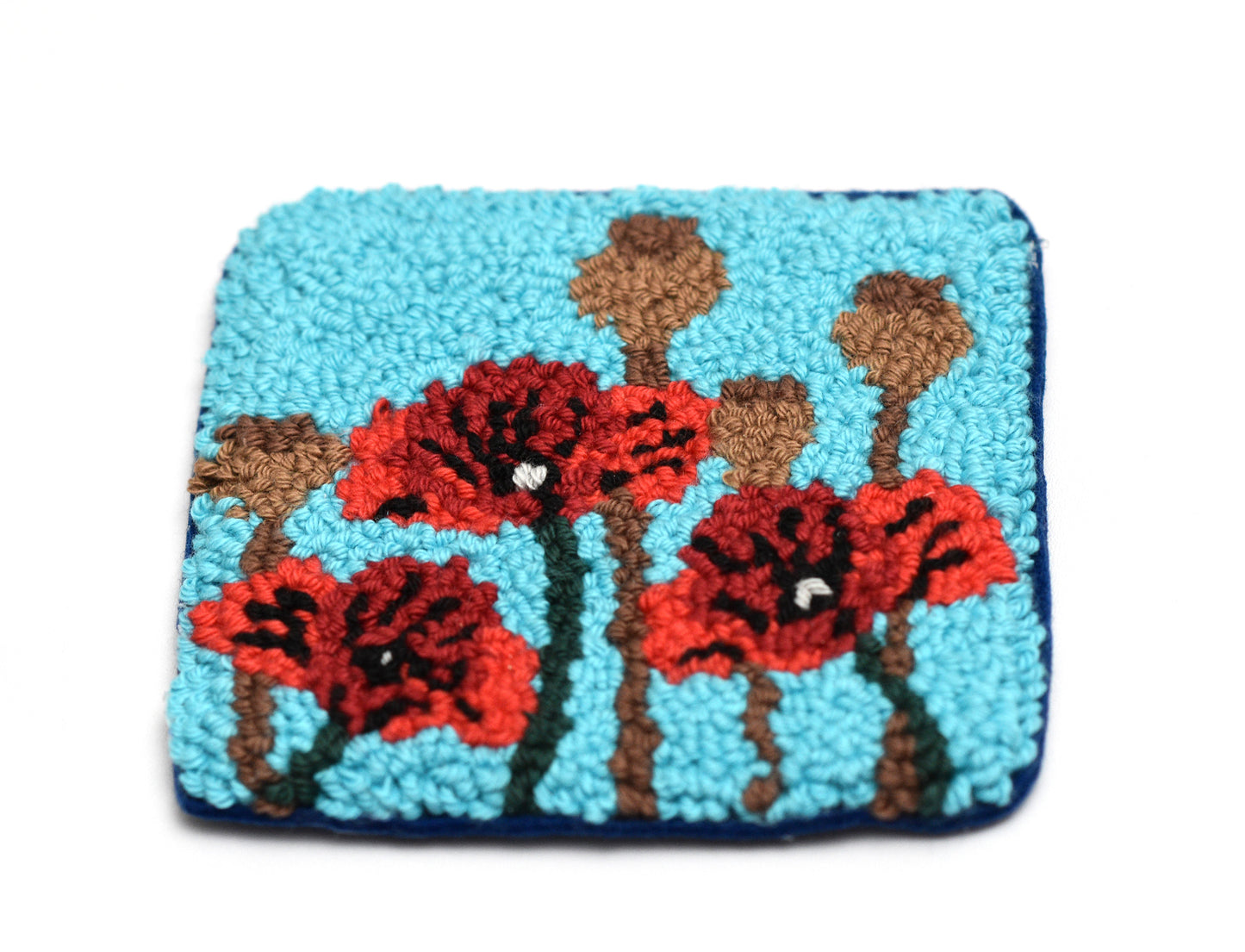 Decorative punch needle coaster with design of red flowers and brown stems on a blue background