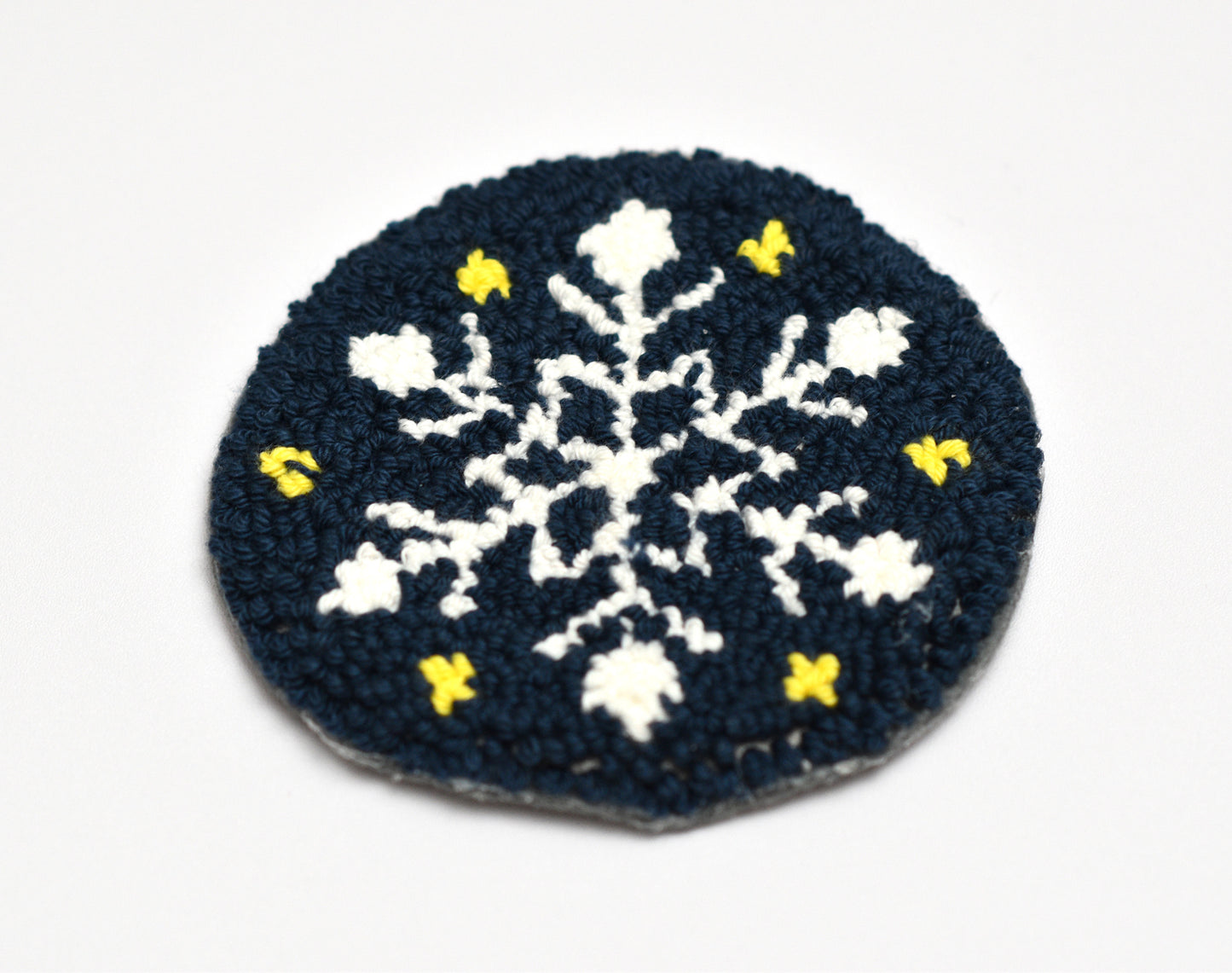 Punch needled snowflake coaster with white snowflakes and yellow accents on a dark blue background