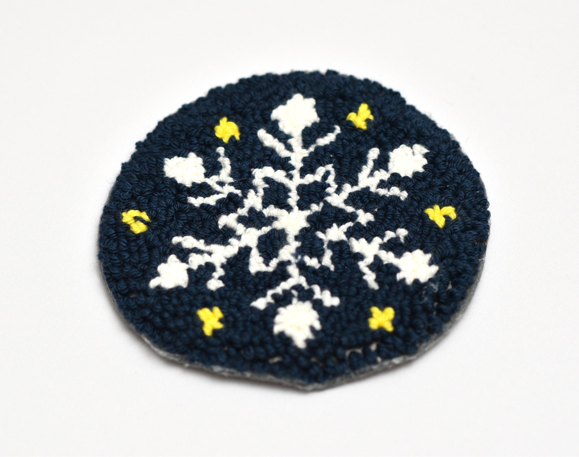 Punch needled snowflake coaster with white snowflakes and yellow accents on a dark blue background