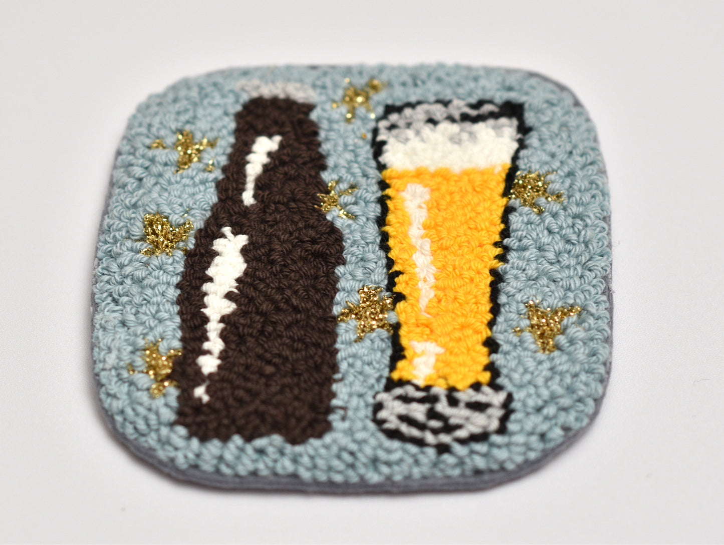 Embroidered coaster with a beer bottle and glass design on a light gray background
