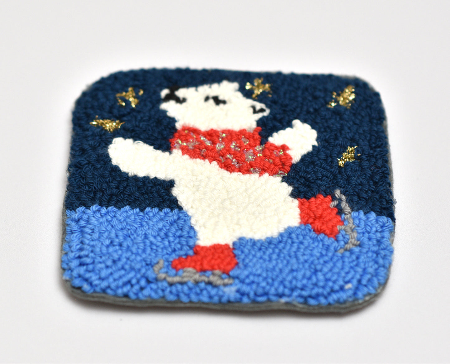 punch needle coaster with a design of a Polar bear in a red scarf on a blue background with gold stars