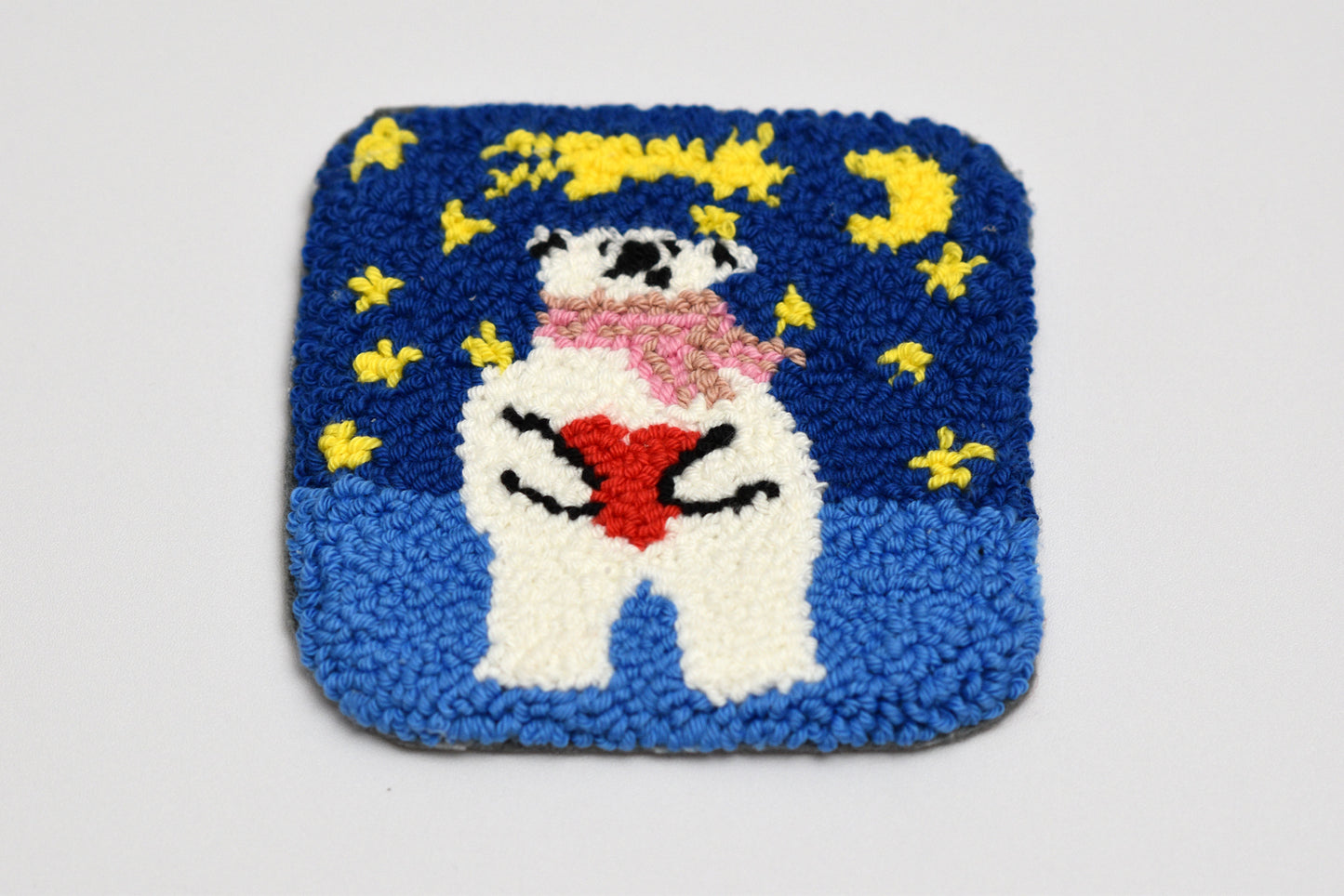 Square punch needled coaster with a bear holding a heart and stars design on a blue background