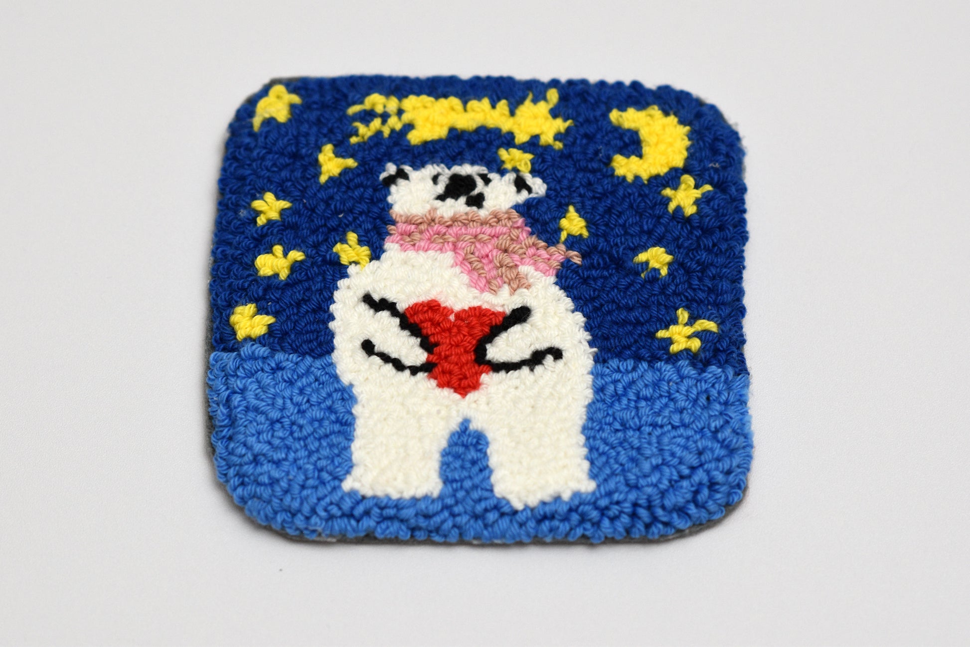 Square punch needled coaster with a bear holding a heart and stars design on a blue background