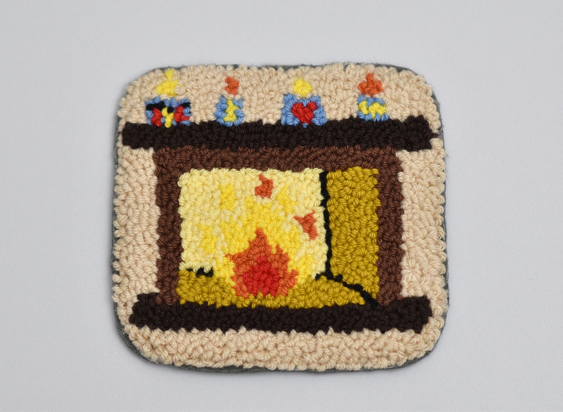 Punch needled coaster with a fireplace design featuring colorful flames and candles on a gray background