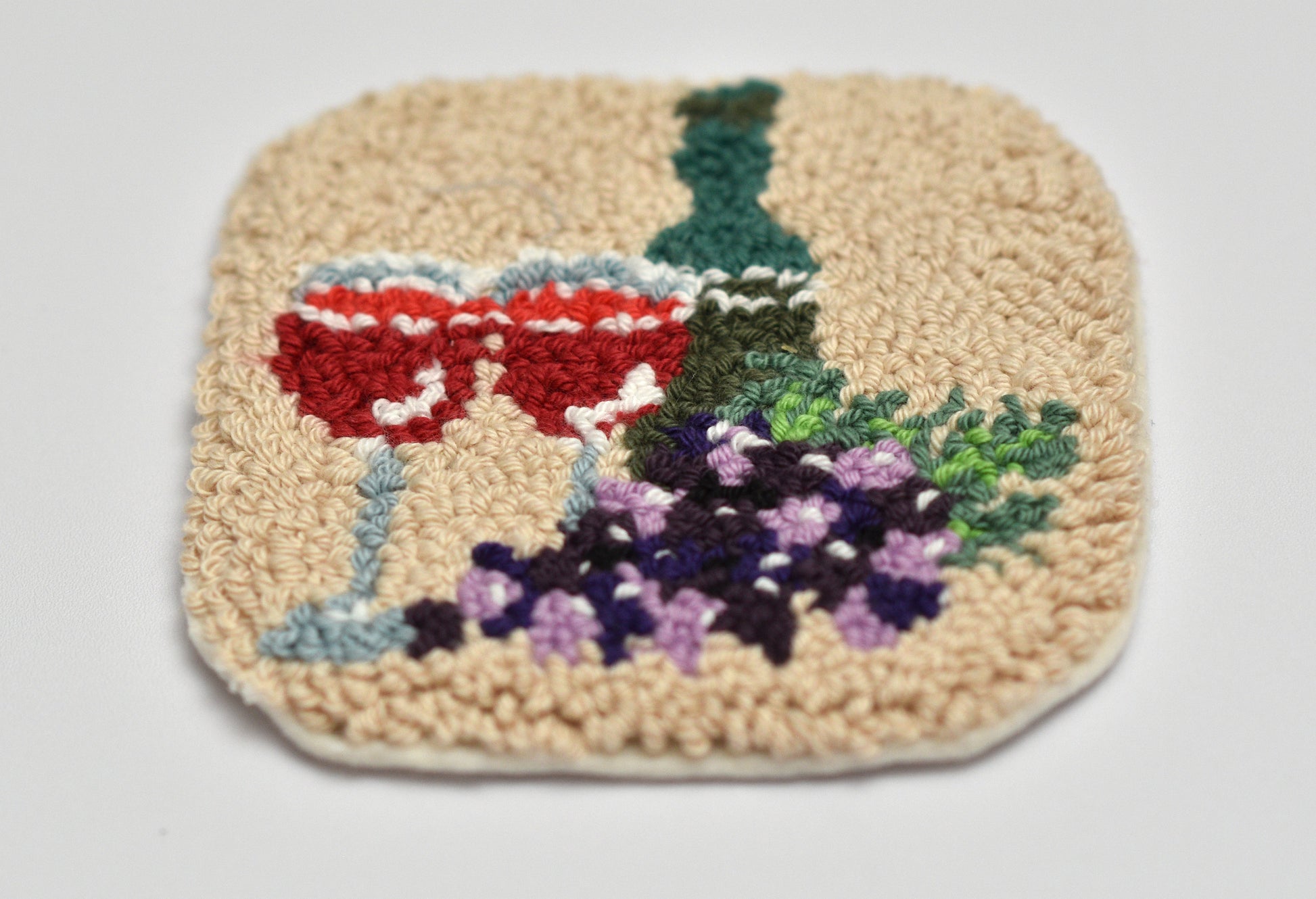 Punch needled coaster with embroidered design of a wine glass and grapes on a plain background