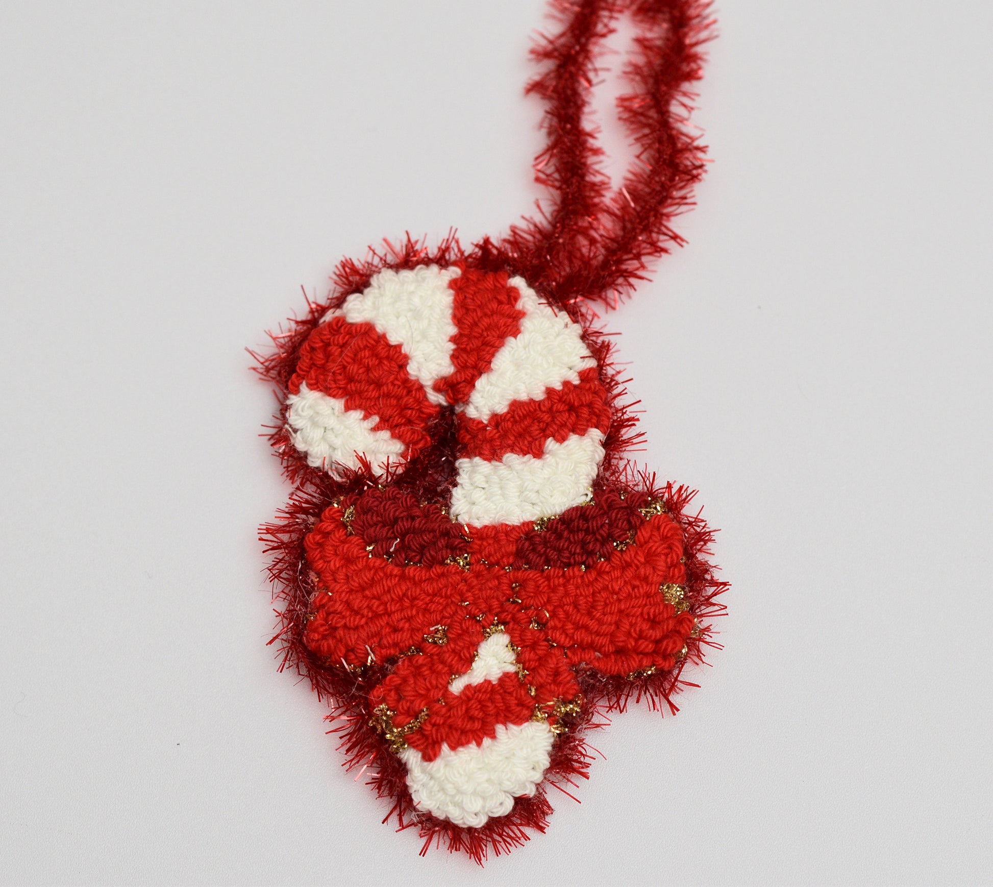 Red and white candy cane punch needle ornament with tinsel on a light gray background