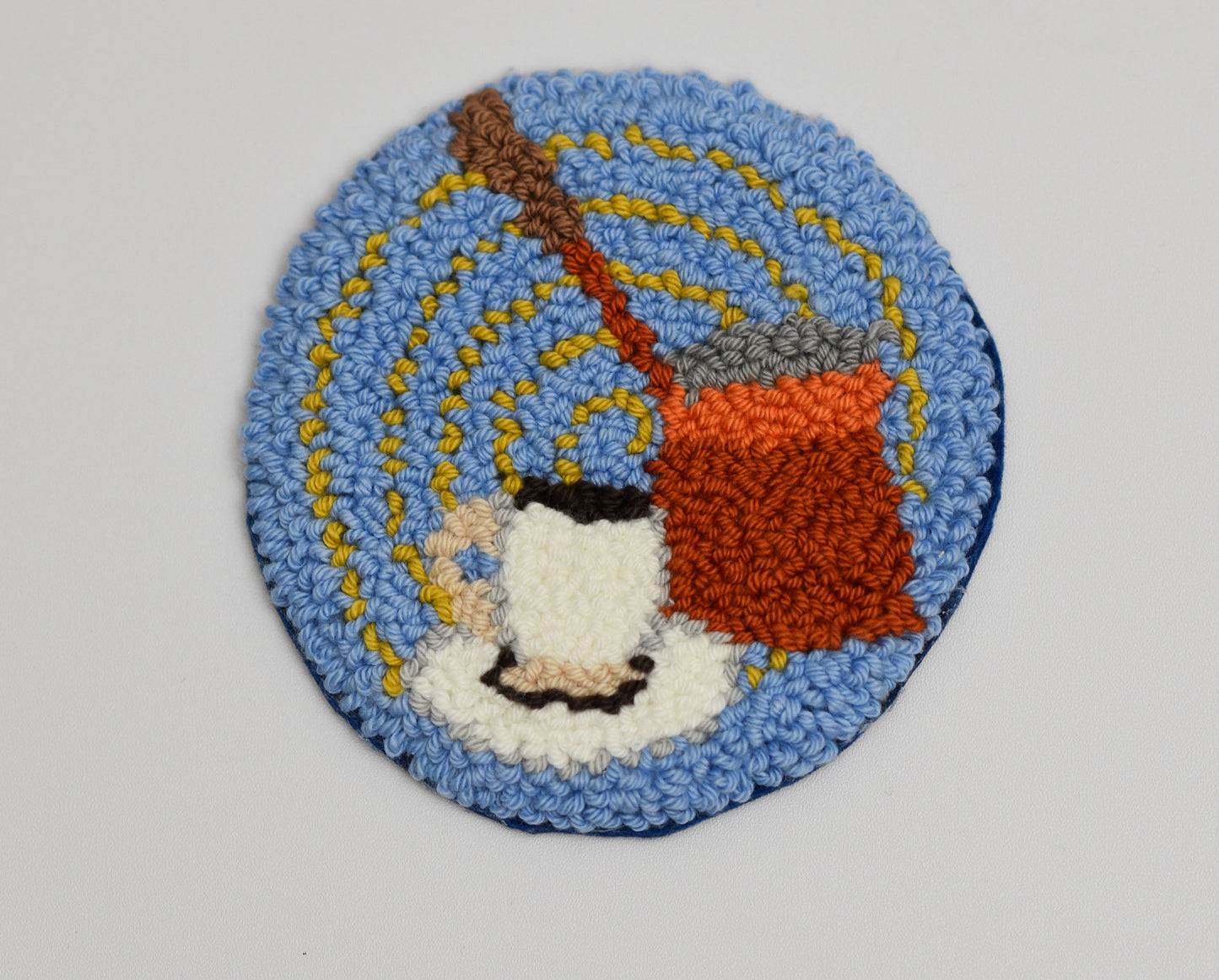 Circular punch needle coaster with a design of a Turkish coffee pot and cup on a blue background