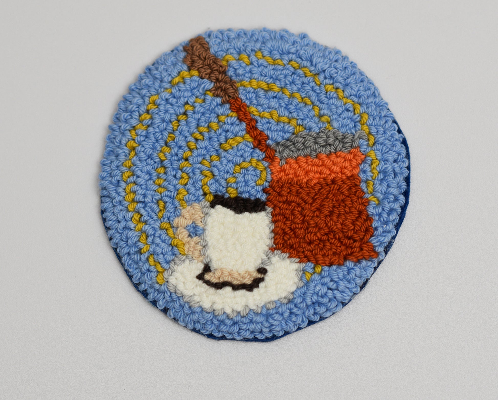 Circular punch needle coaster with a design of a Turkish coffee pot and cup on a blue background