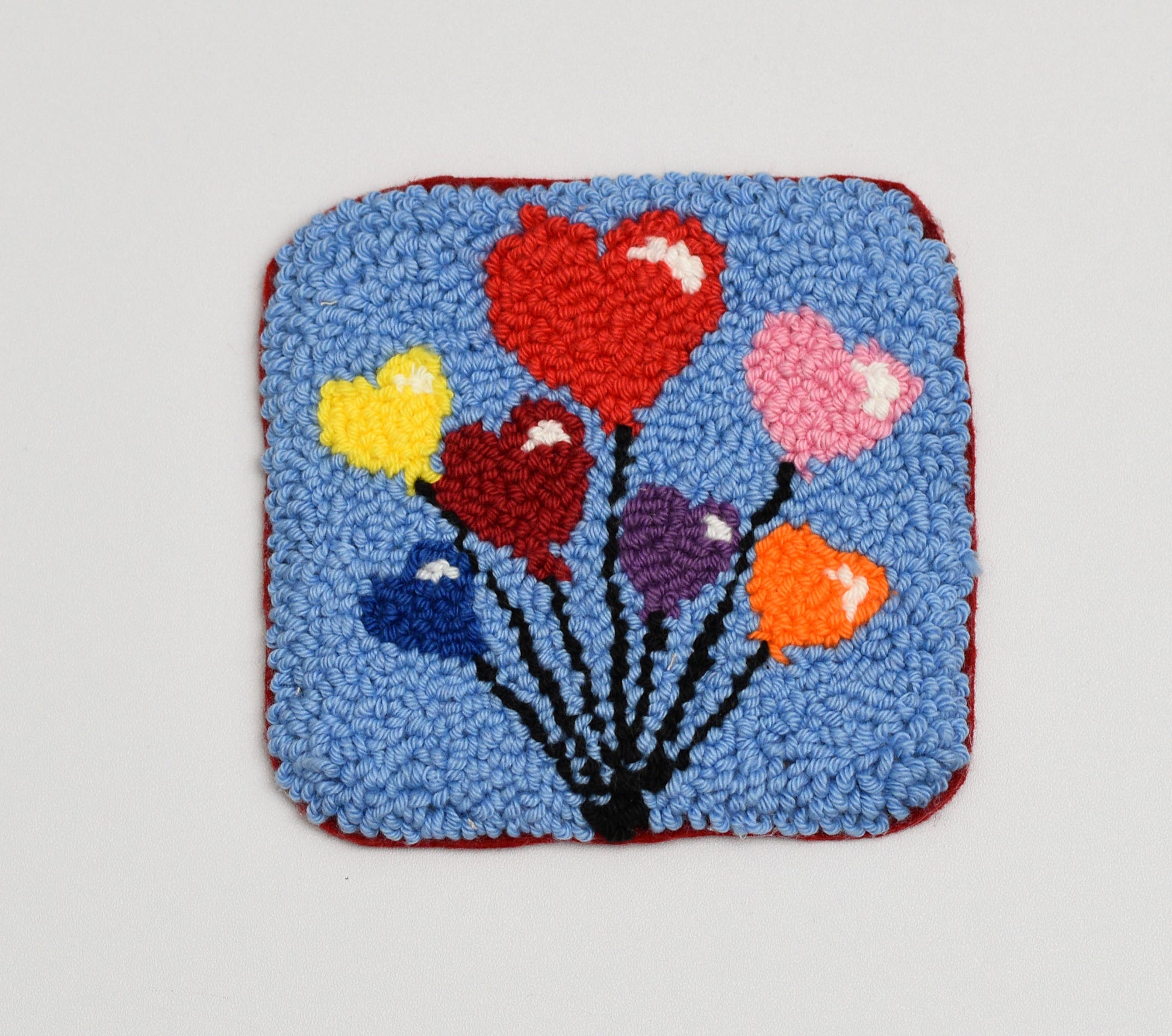 Square punch needle coaster with colorful heart-shaped balloons on a blue background