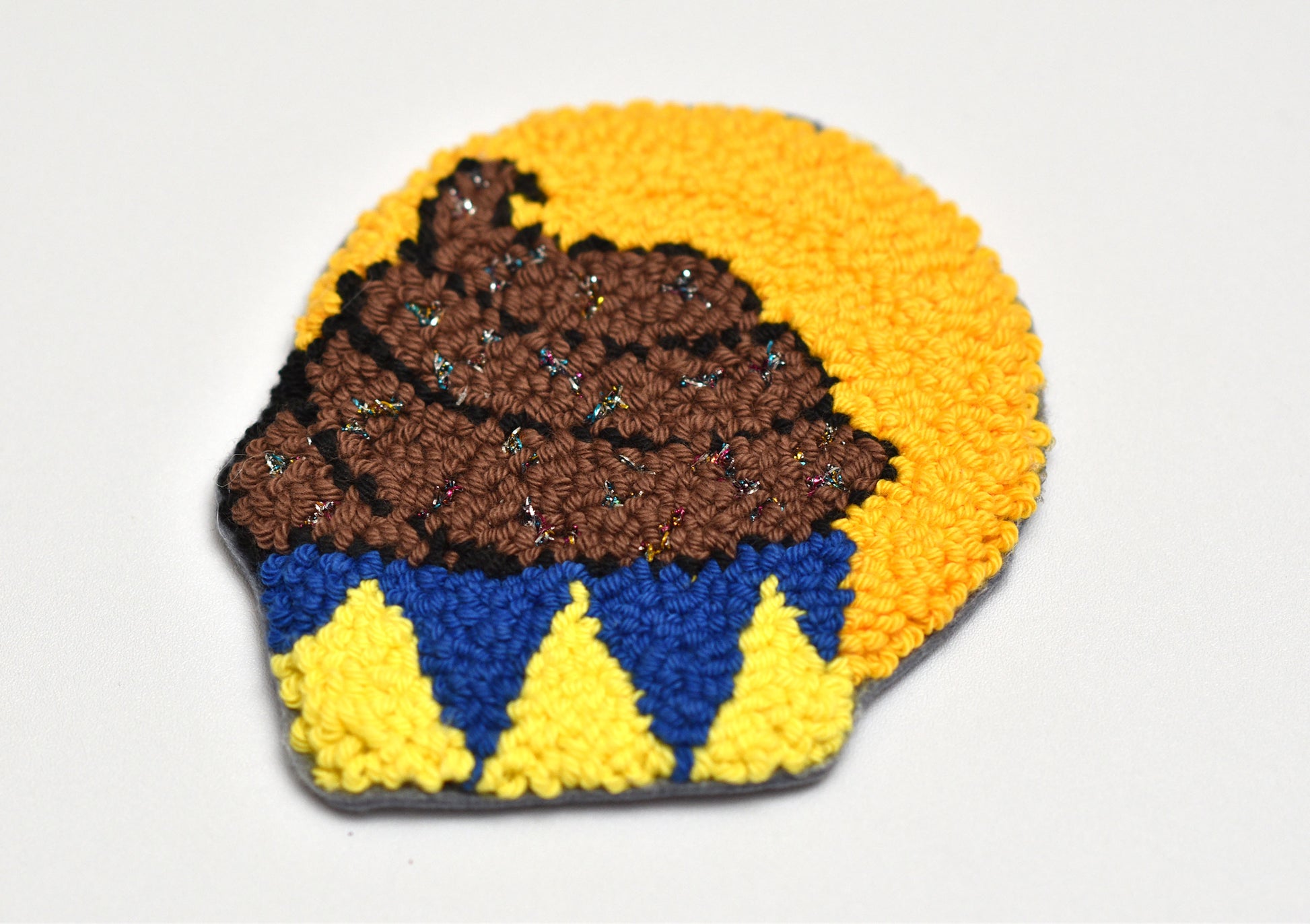 Punch needle coaster shaped as a cupcake with brown top, blue middle, and yellow bottom on a white background