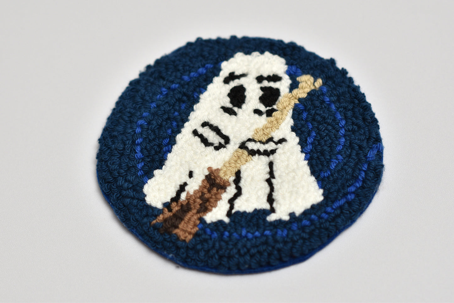 Round punch needle coaster with a ghost design holding a broom on a blue background