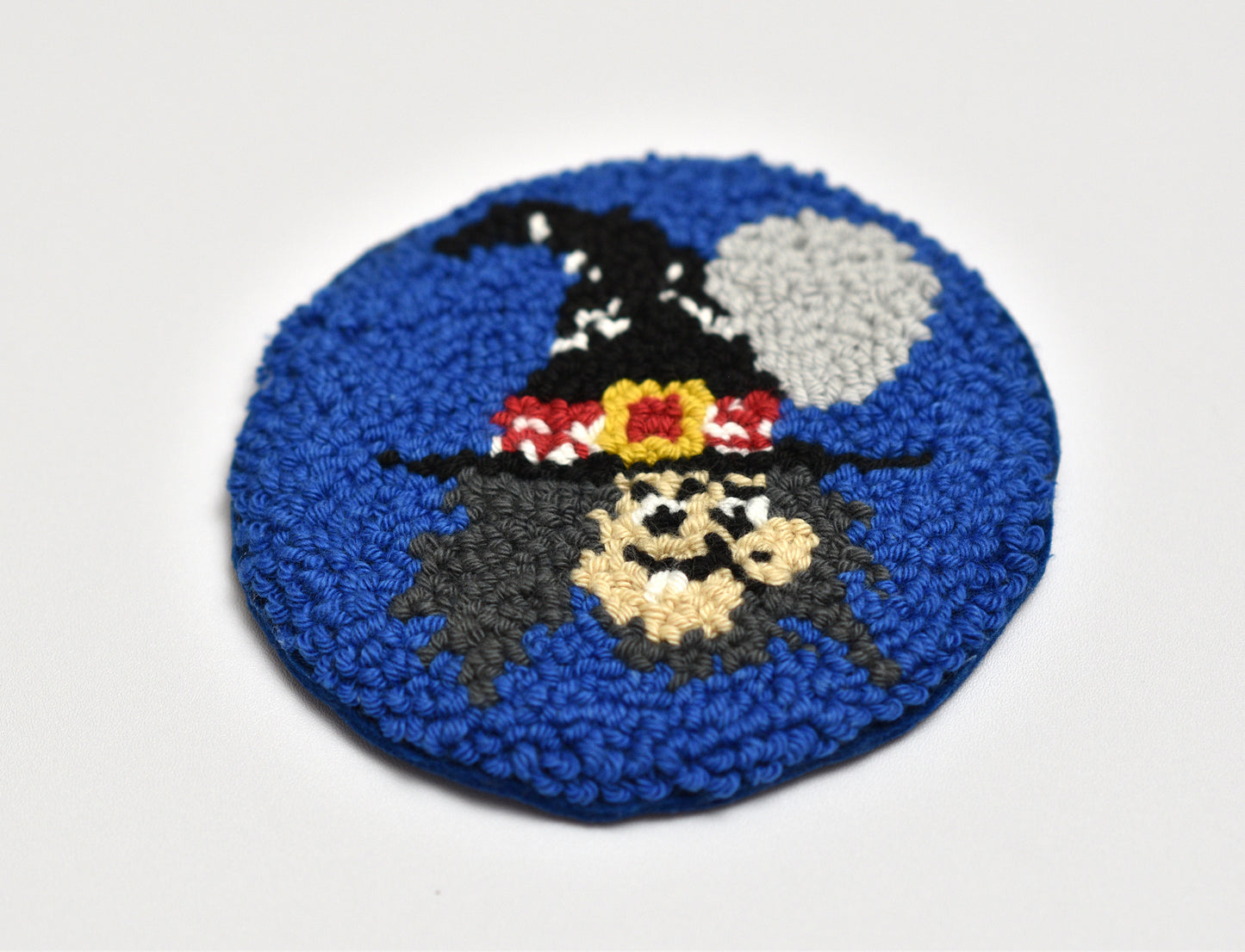 Round blue punch needle coaster with a witch design on a white background