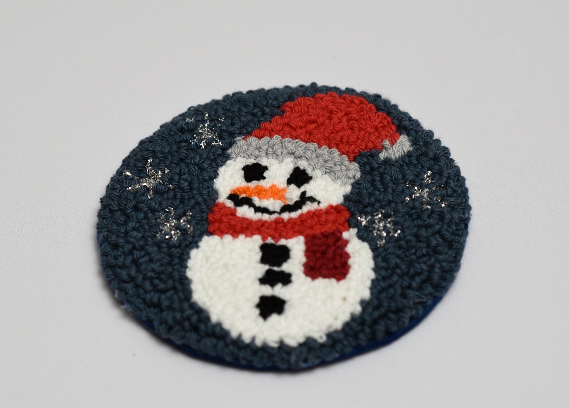 Round punch needled coaster with a snowman design on a gray background