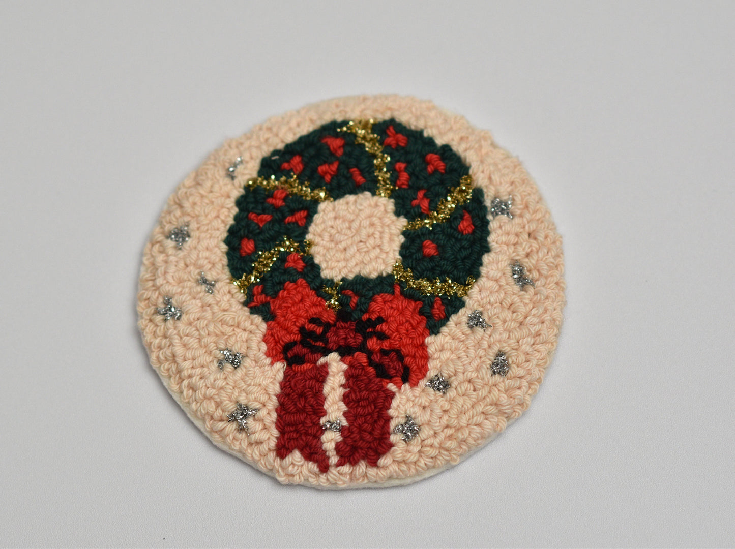 Round punch needle coaster with a Christmas wreath design on a gray background