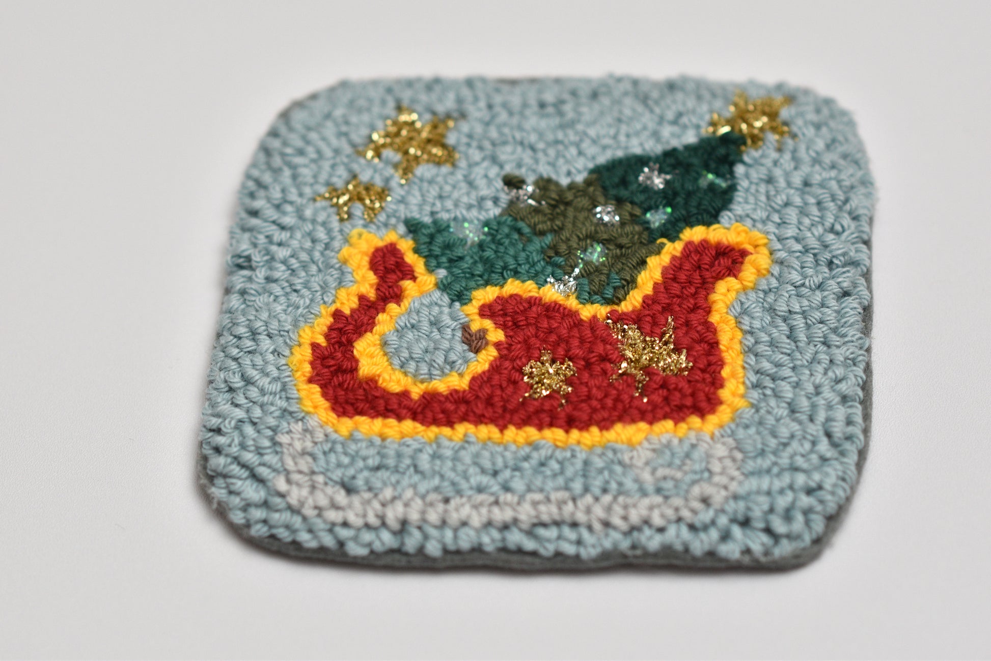 Decorative punch needle mug rug with a Christmas scene featuring a sleigh, tree, and stars on a light blue background.