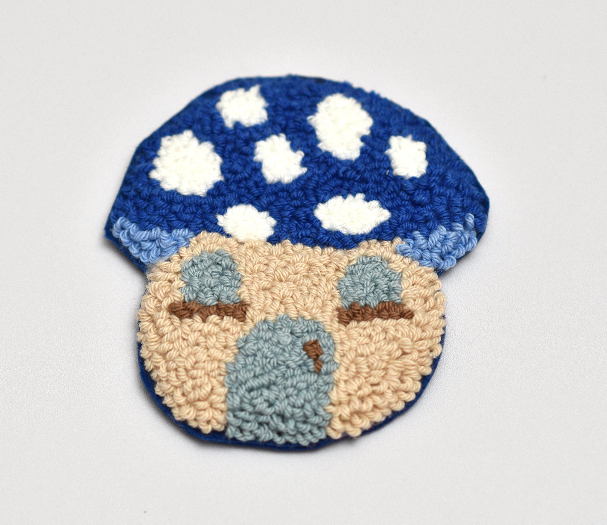 Punch needled mushroom house shaped coaster with blue cap and beige body on a light gray background