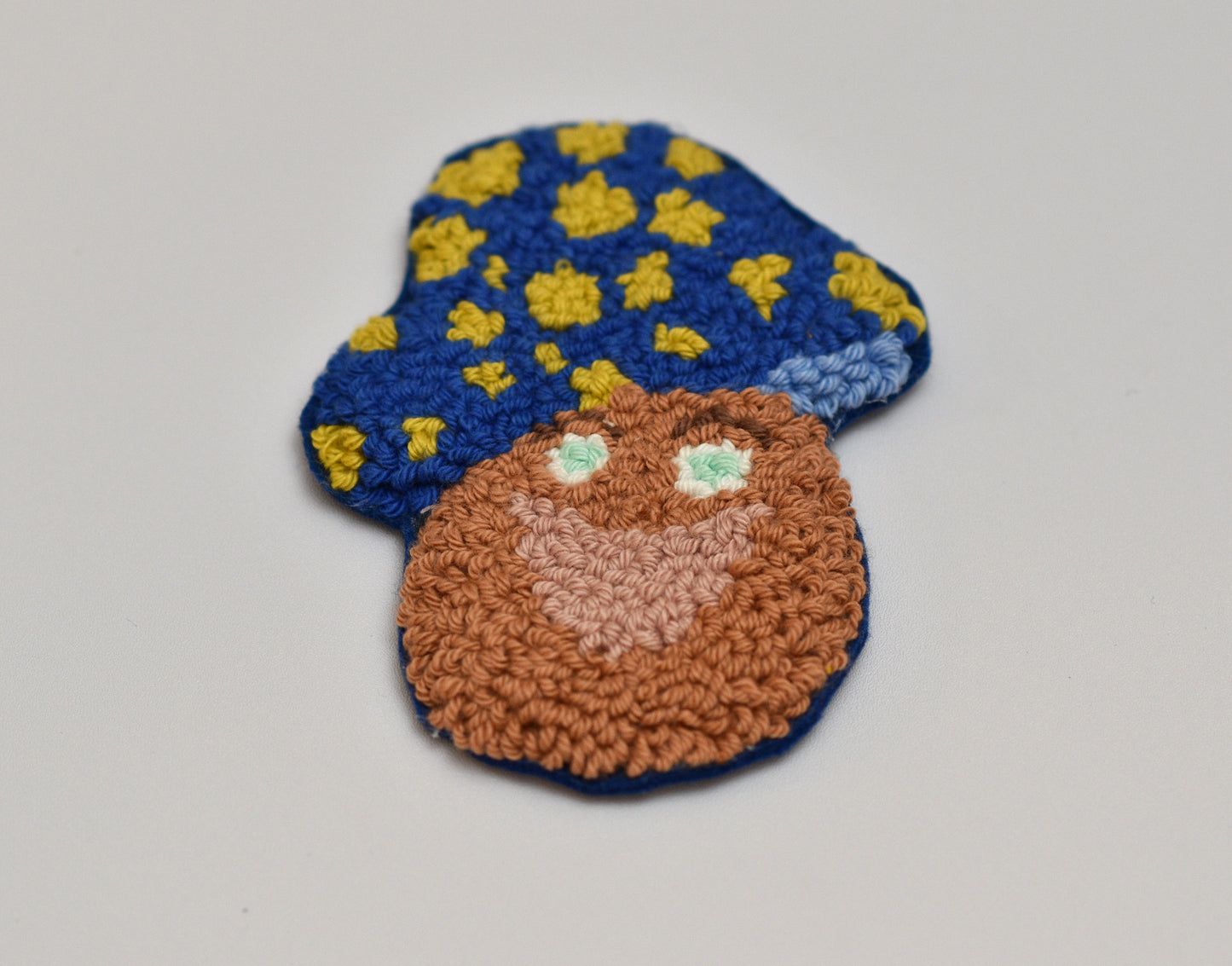 Punch needled mushroom designed coaster with blue cap and brown body on a light gray background