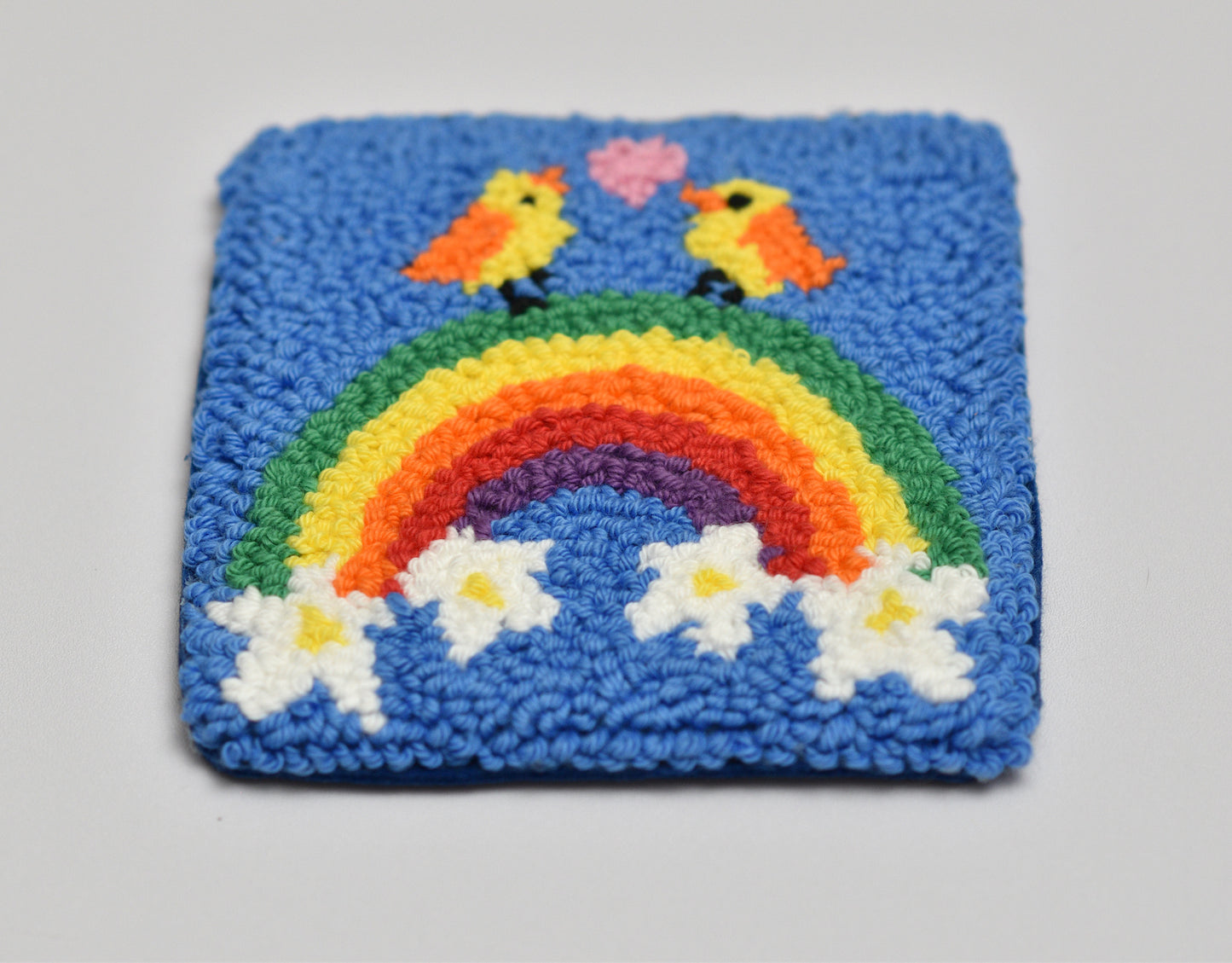 Punch needled square coaster with a rainbow, birds, and flowers on a blue background