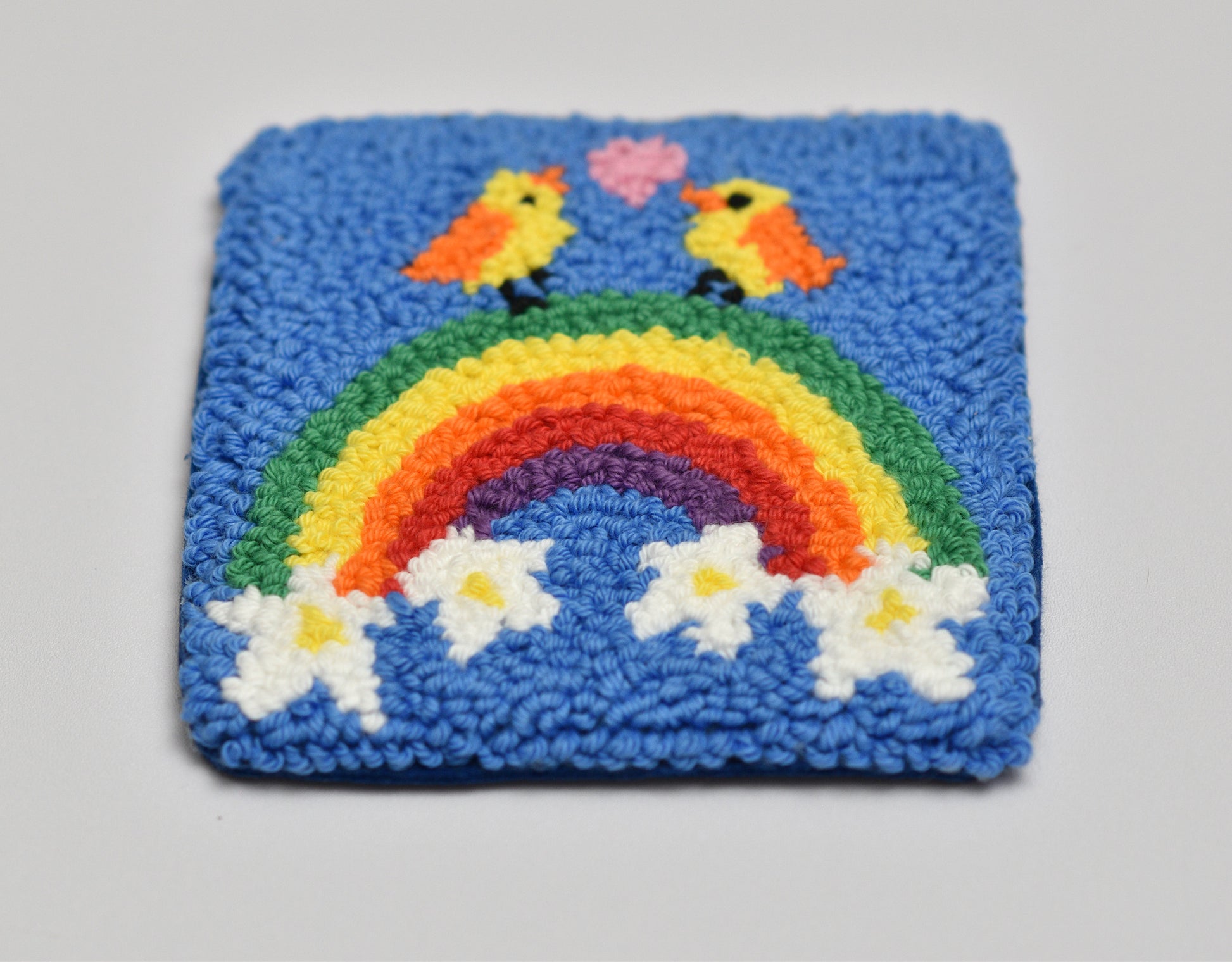 Punch needled square coaster with a rainbow, birds, and flowers on a blue background