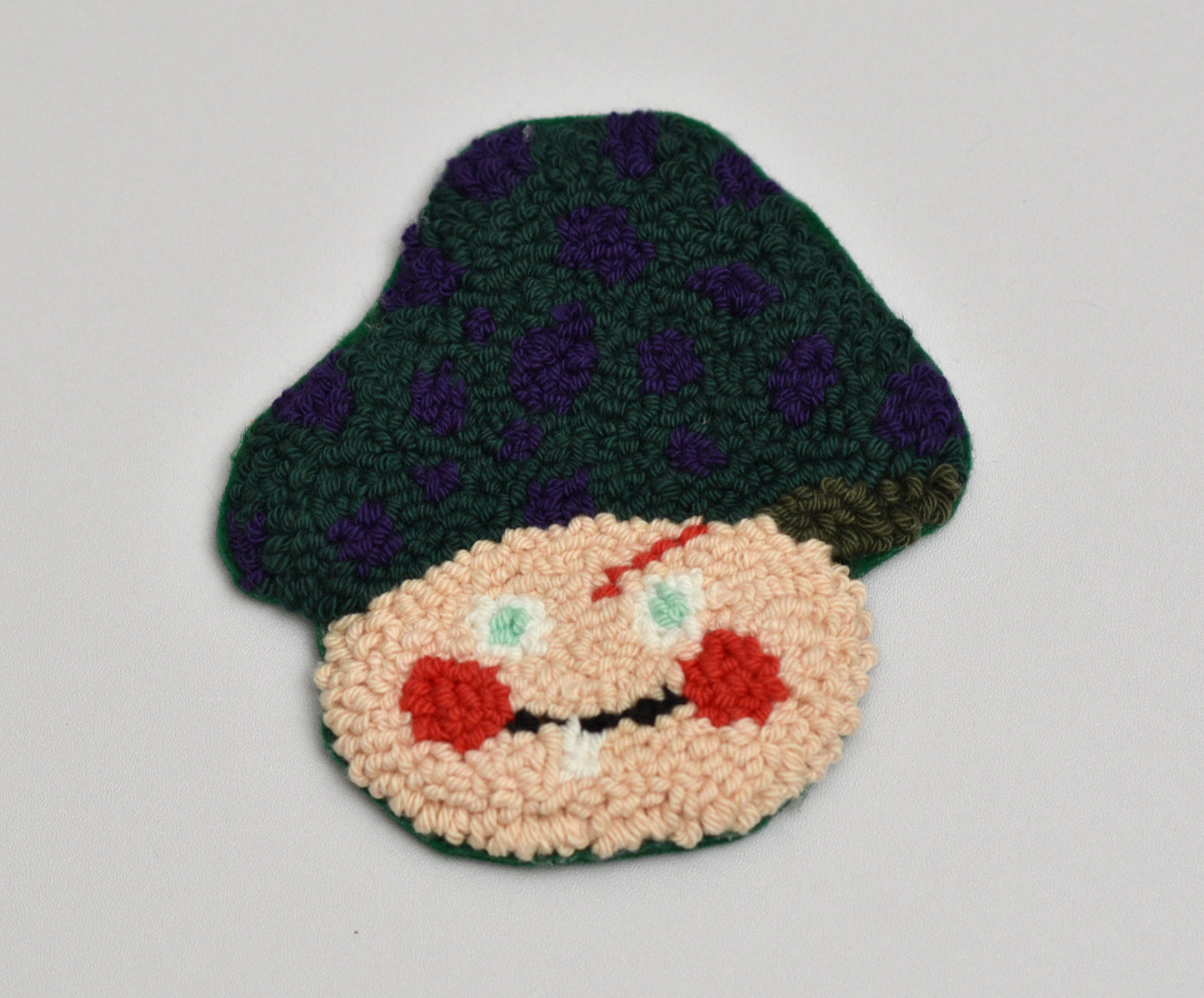 Punch needled coaster with design of a mushroom face with a green hat on a light gray background