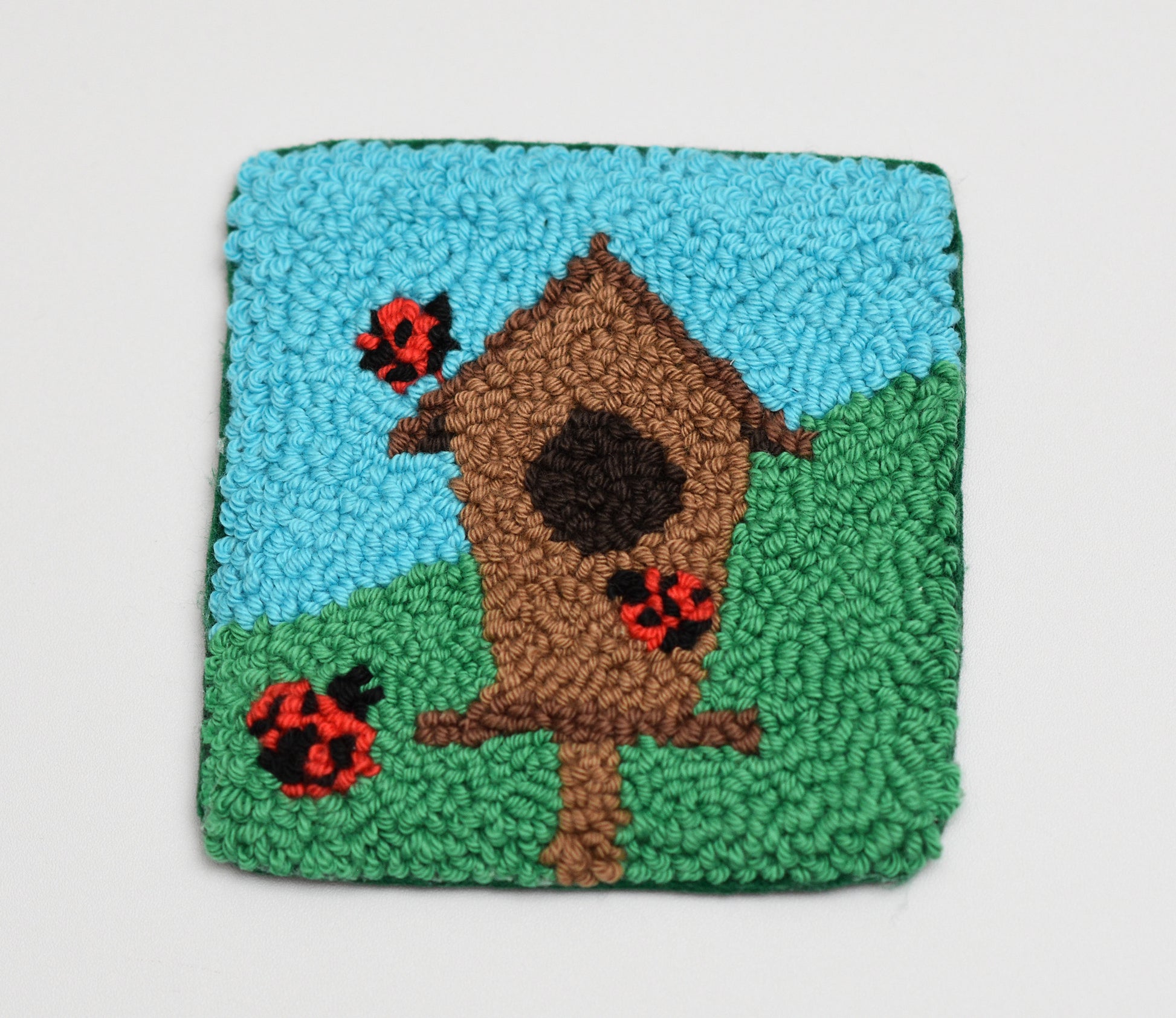 Textured square coaster with a birdhouse and ladybugs on a light gray background