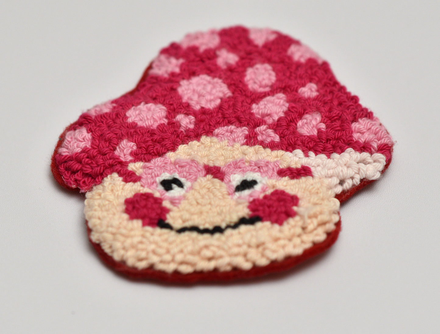 Handmade punch needle coaster with mushroom character with a red hat and pink polka dots on a light gray background