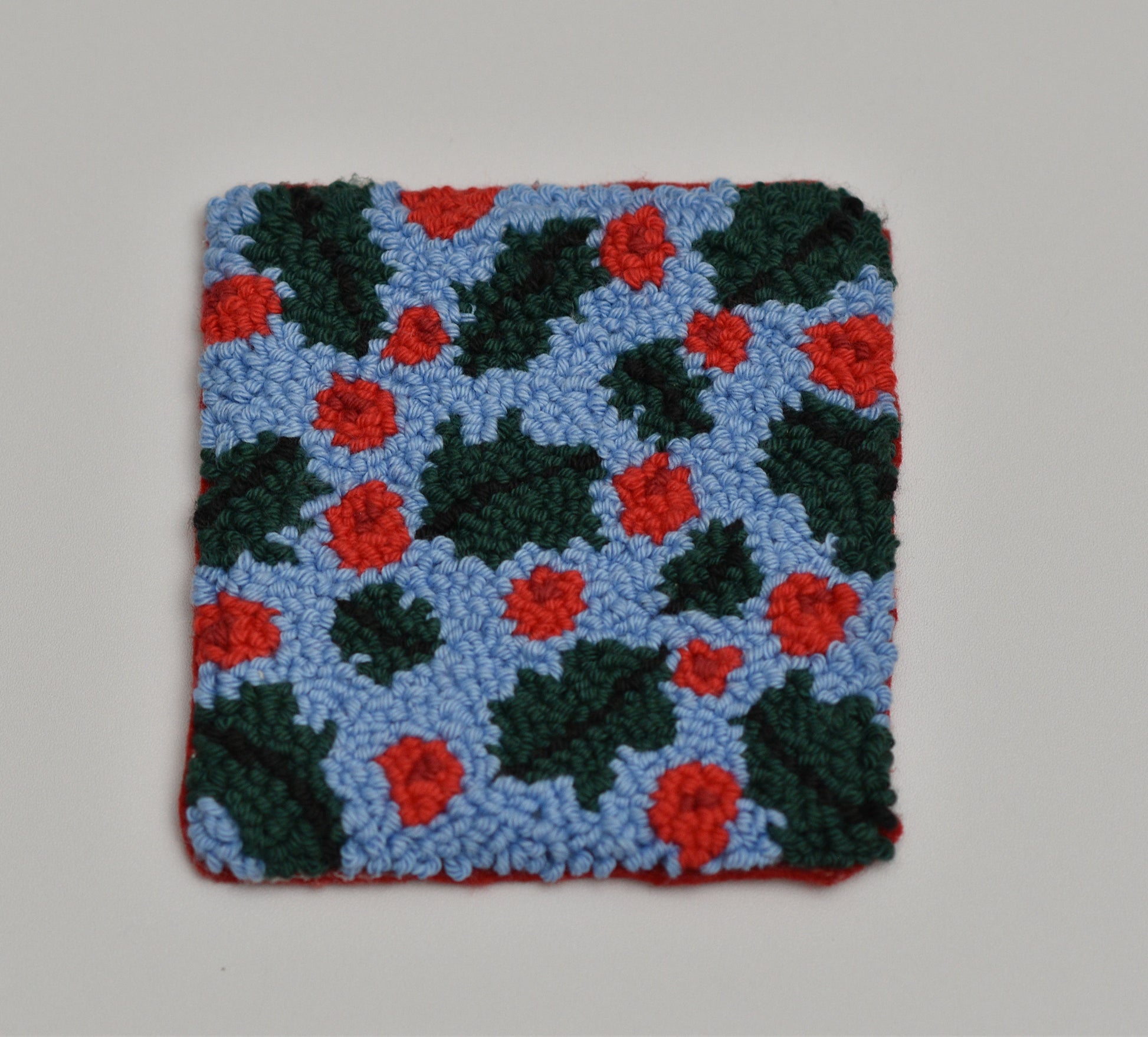 Square punch needle coaster with mistletoe  pattern featuring red flowers on a blue background