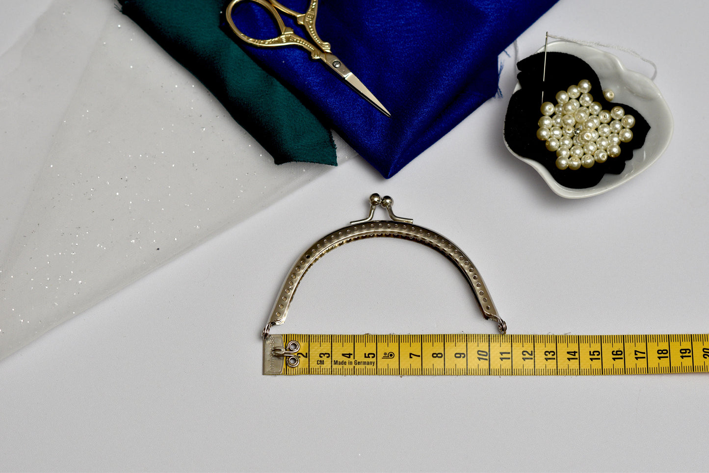 Metallic purse frame with a measuring tape for scale, on a white surface with a blue fabric and pearl container in the background.