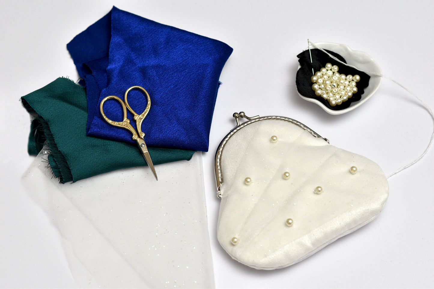 Heart-shaped white purse with pearl embellishments, blue fabric, green fabric, and gold scissors on a white background.