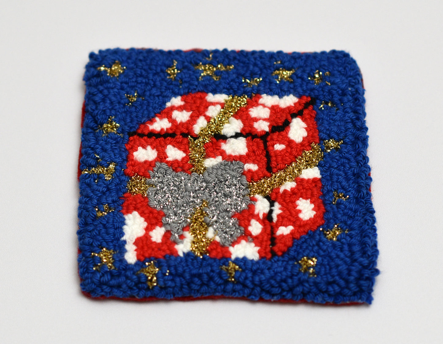 Decorative punch needle coaster with red gift package, gray bow, and gold accents on a blue background.