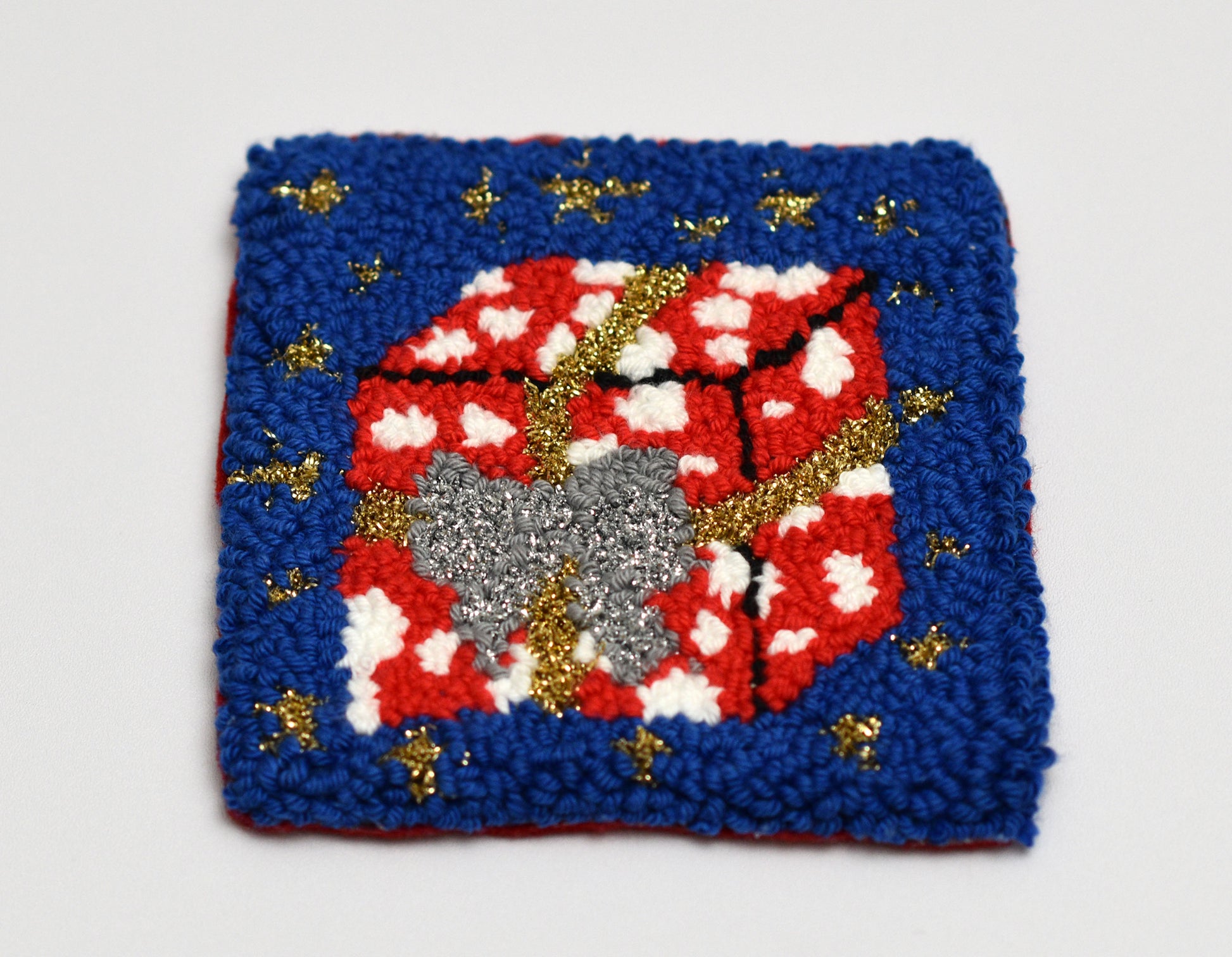 Decorative punch needle coaster with red gift package, gray bow, and gold accents on a blue background.