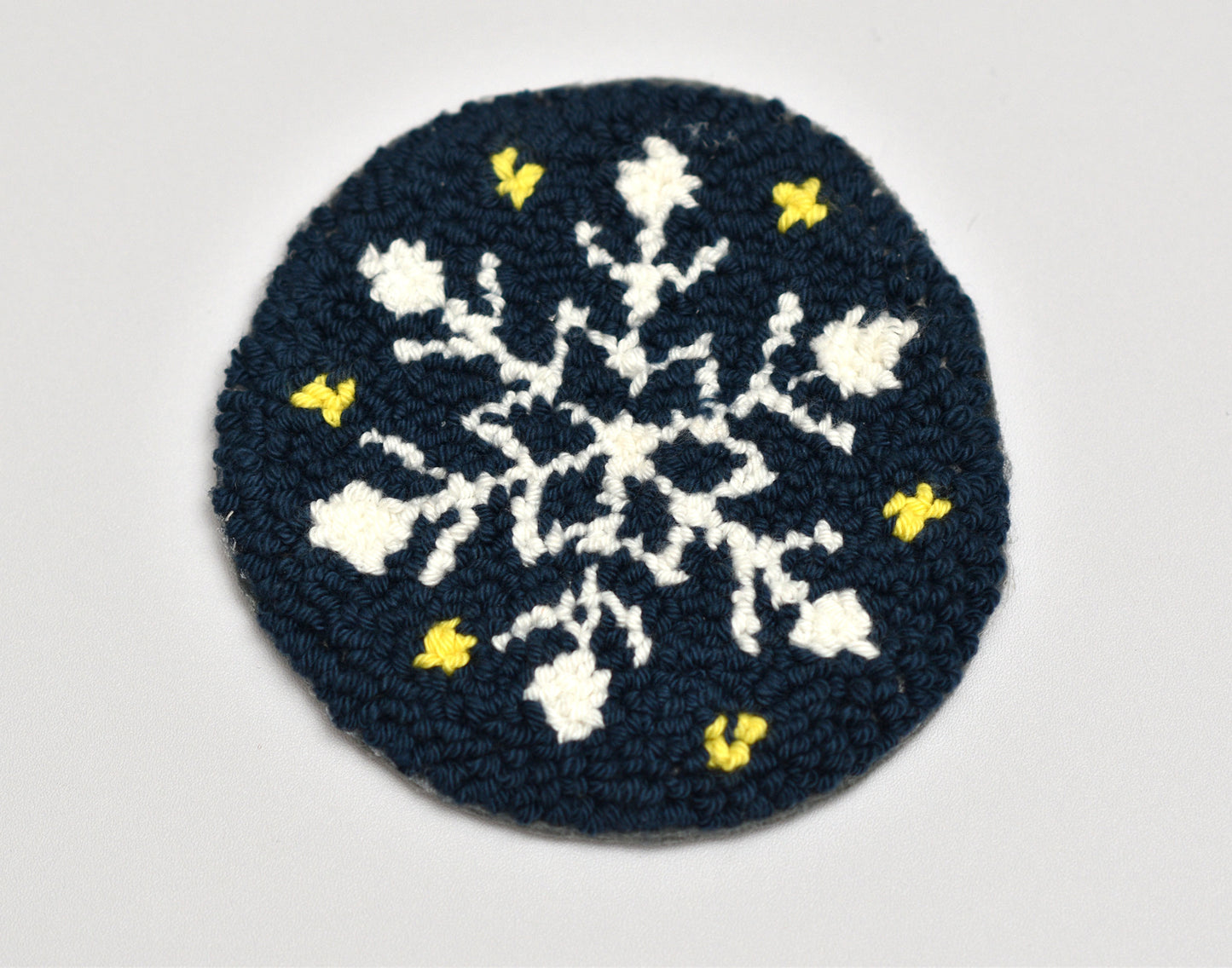 Dark blue punch needled coaster with white snowflake pattern and yellow accents on a light gray background