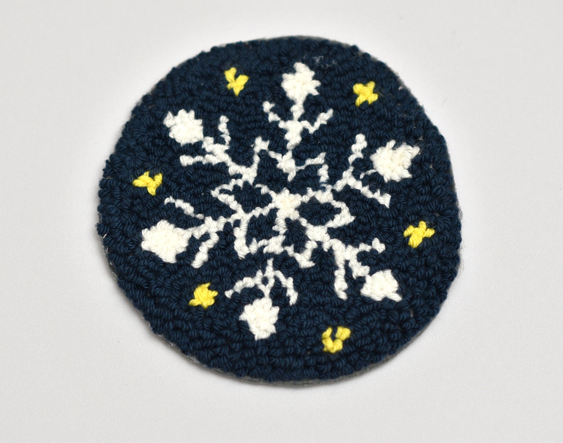 Dark blue punch needled coaster with white snowflake pattern and yellow accents on a light gray background