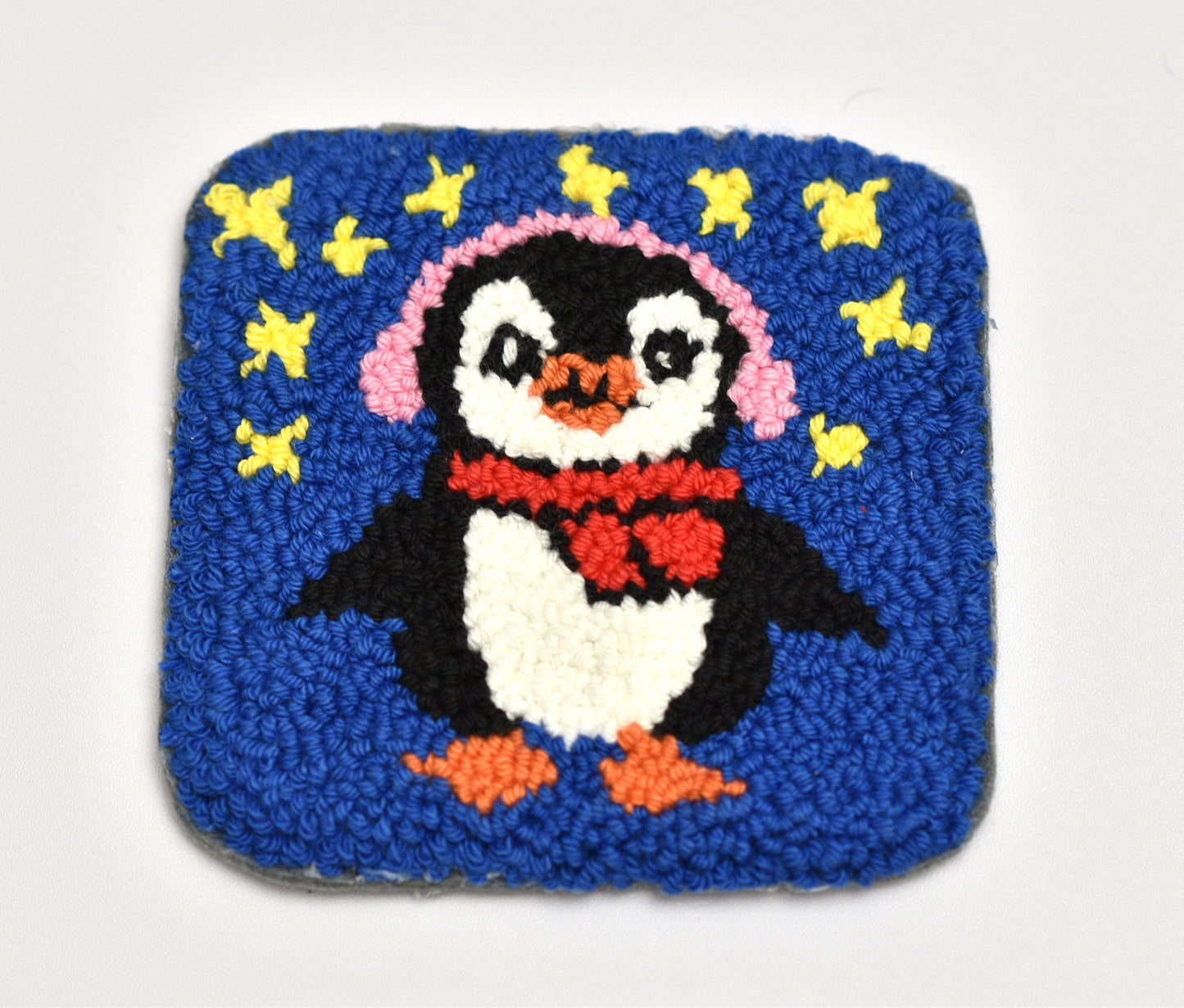 Embroidered coaster of a penguin wearing a pink headphones and red scarf on a blue background with yellow stars.