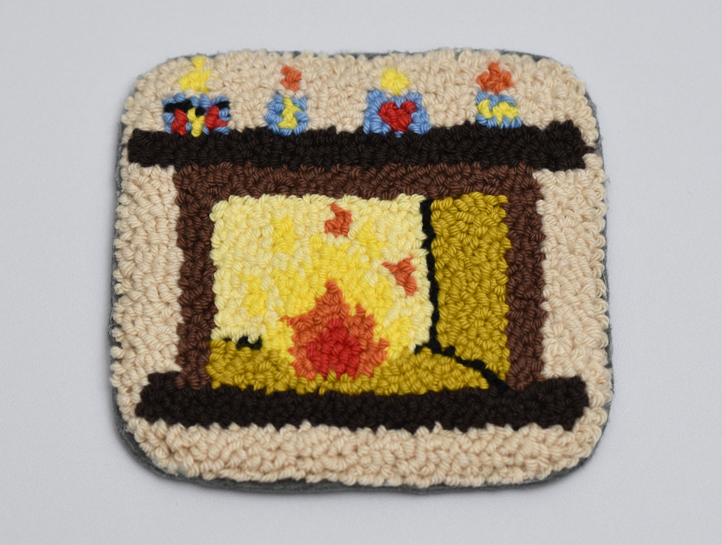 Punch needled square coaster with fireplace design and colorful candles on a gray background