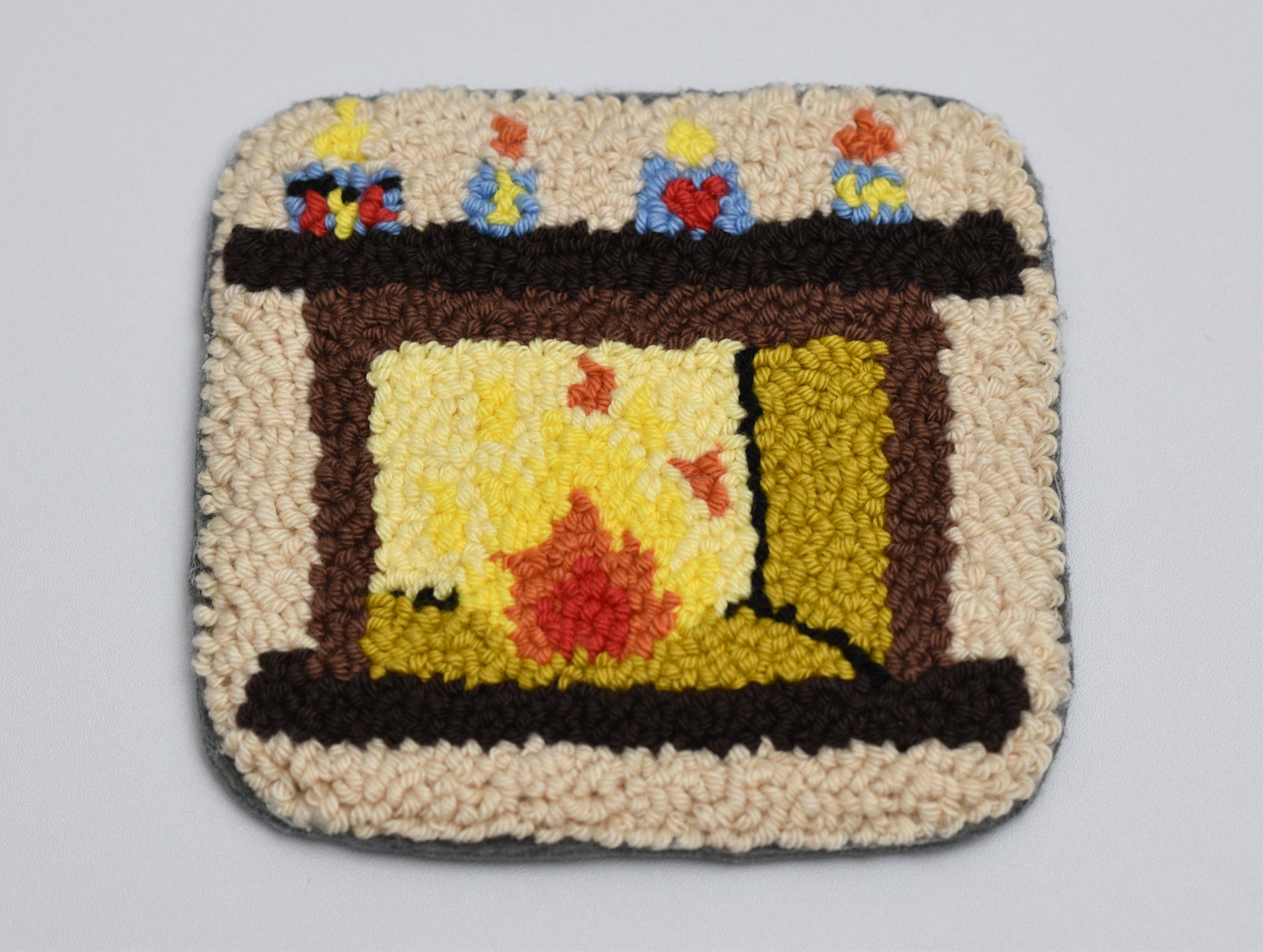 Punch needled square coaster with fireplace design and colorful candles on a gray background