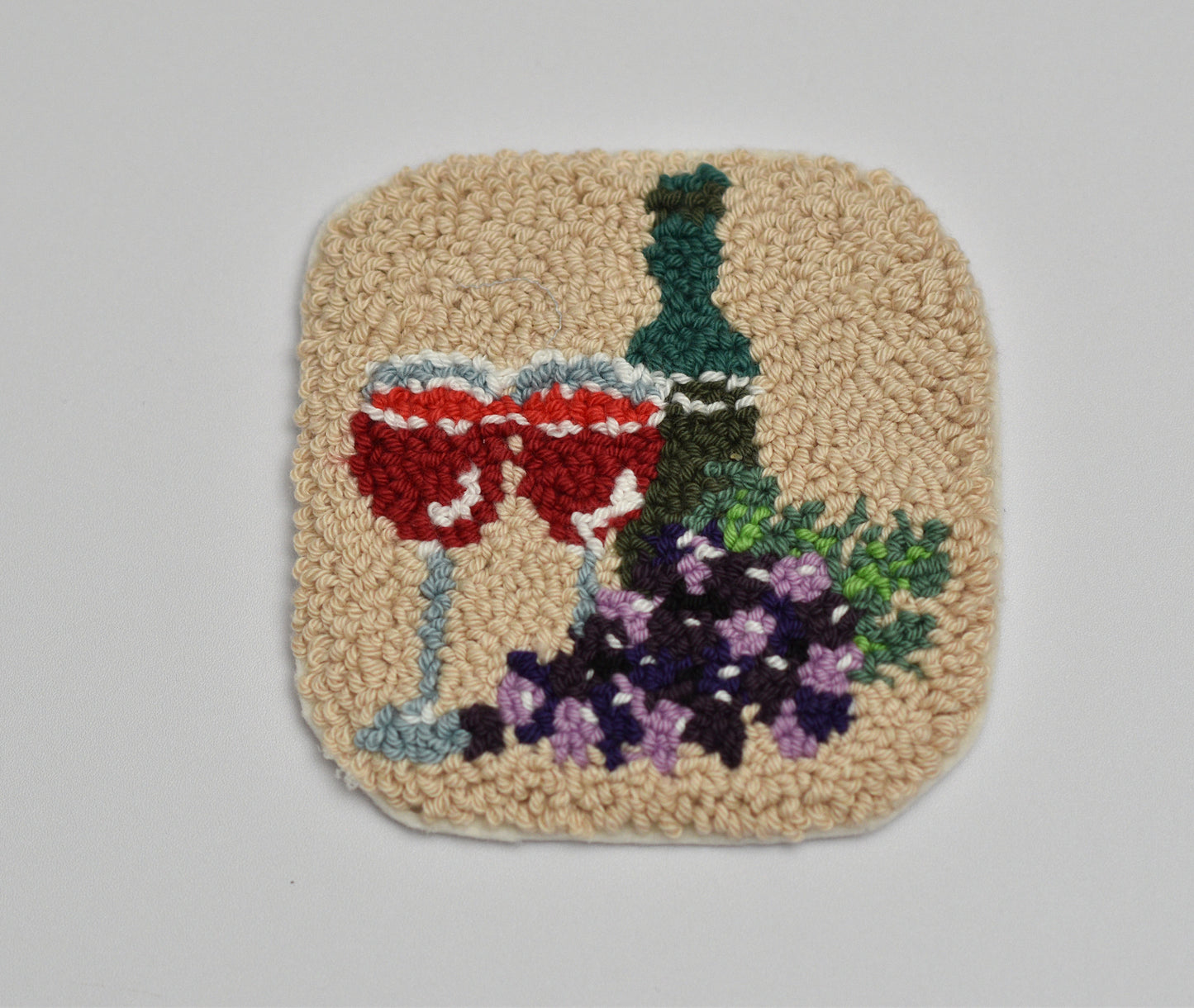 Punch needled coaster with embroidered design of a wine glass, bottle, and grapes on a beige background