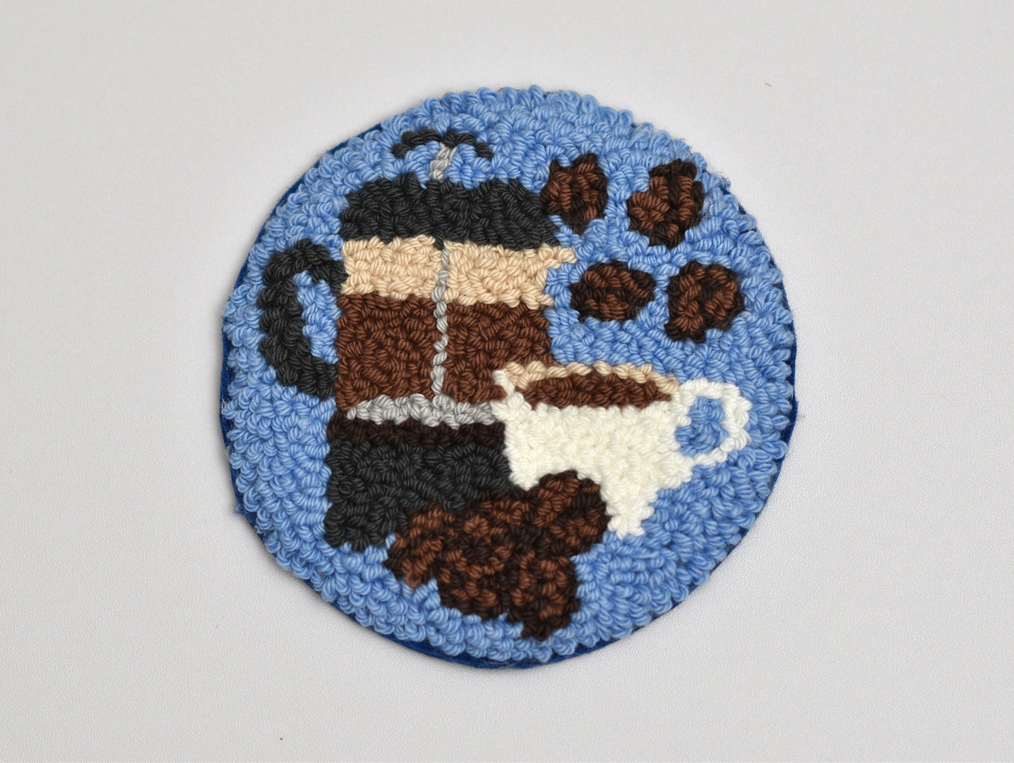 Round woven punch needle coaster with coffee-themed design on a light gray background