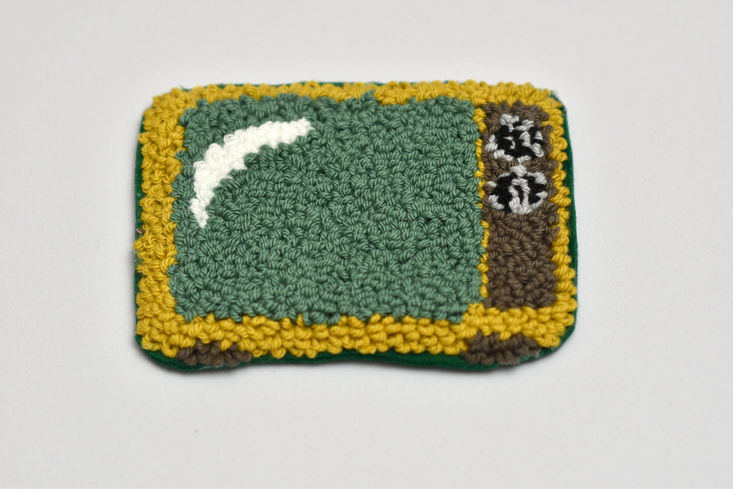Punch needle coaster with retro TV design on green center, yellow border, and white and black designs on a light gray background