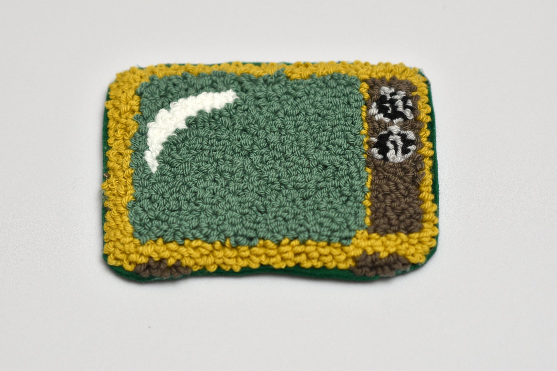 Punch needle coaster with retro TV design on green center, yellow border, and white and black designs on a light gray background
