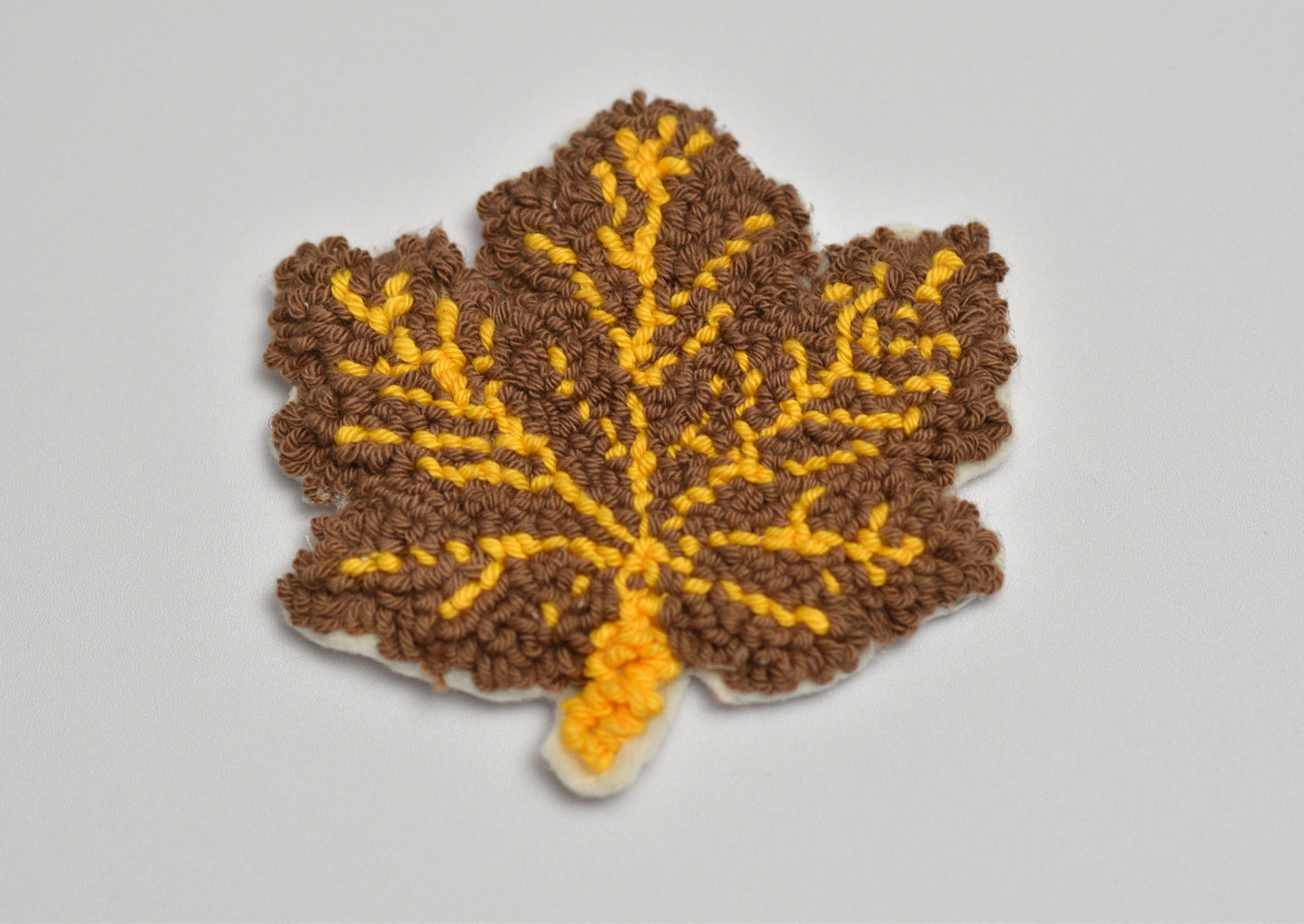 Embroidered leaf-shaped coaster with brown and yellow pattern on a light gray background