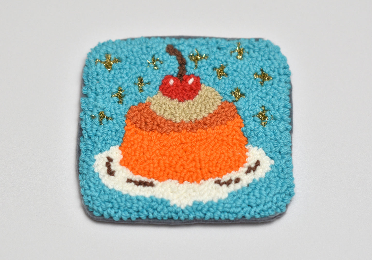 Embroidered coaster with a cake design featuring a cherry on top, on a light gray background.