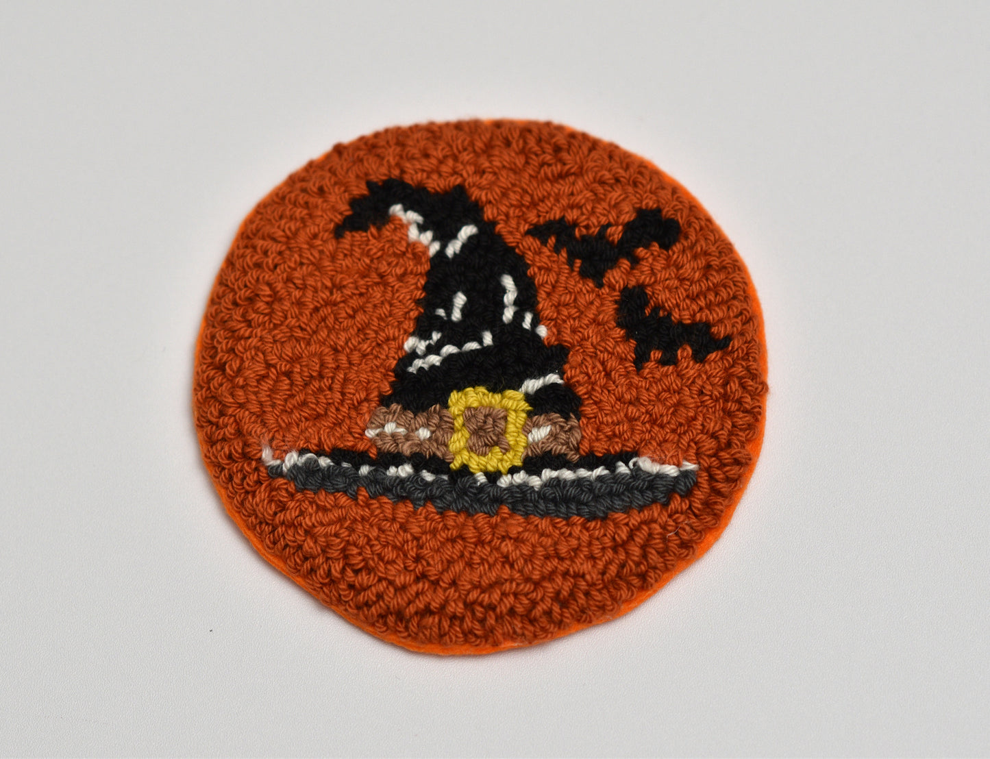 Round punch needle coaster with a witch hat design on an orange background