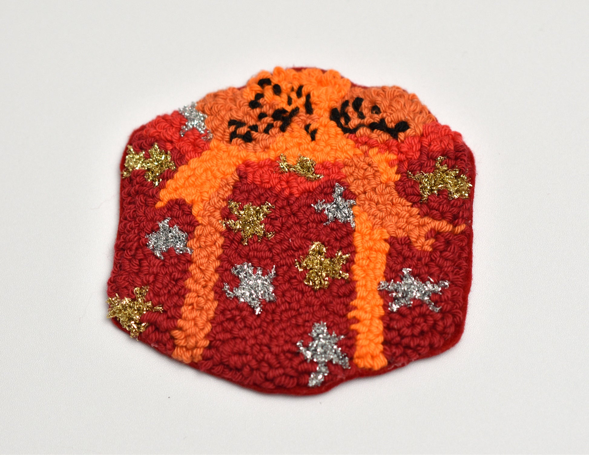 Gift box shaped punch needle coaster with orange center and red border featuring decorative elements on a light gray background