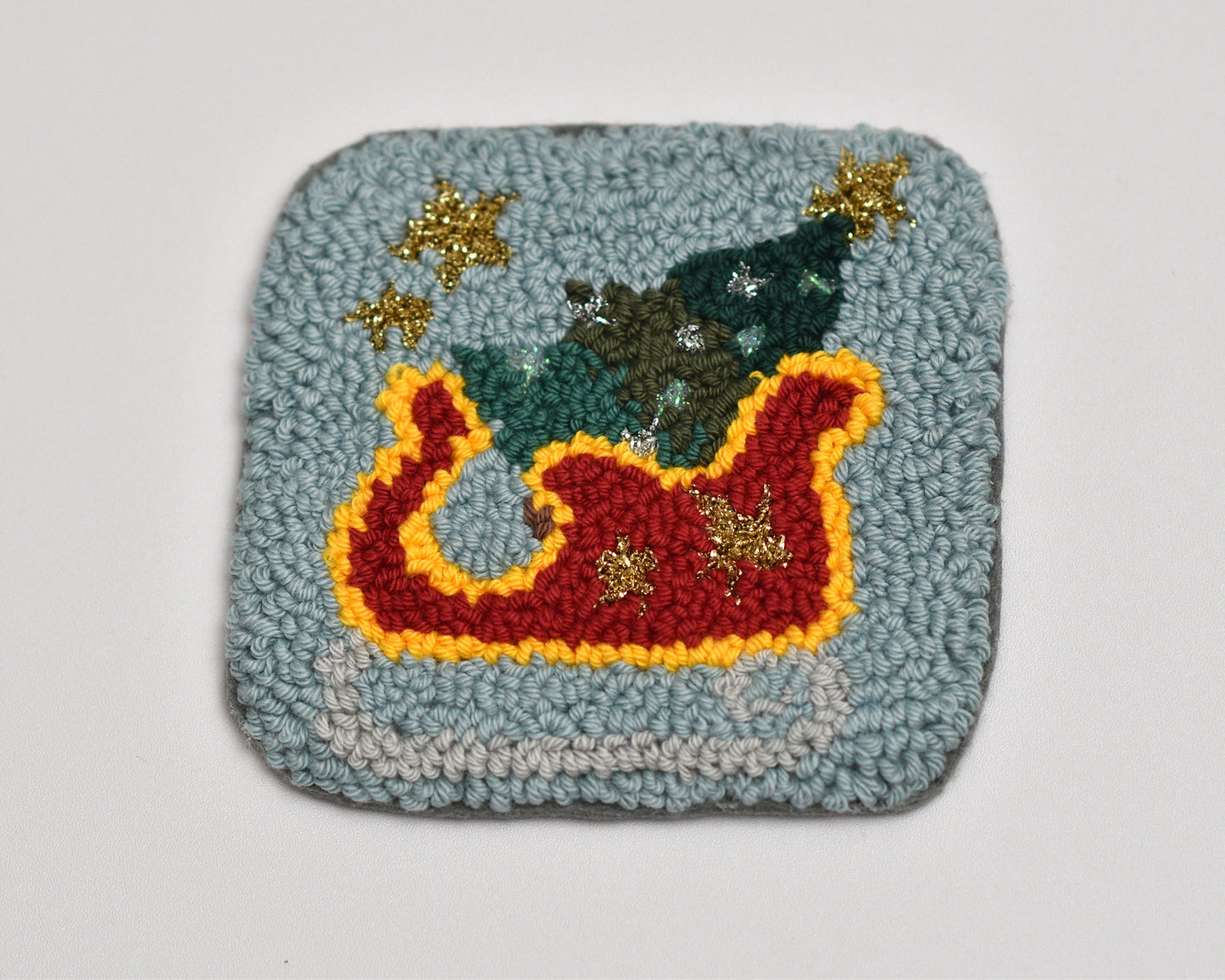 Textured square punch needle coaster with a colorful design of a sleigh, trees, and stars on a light gray background.